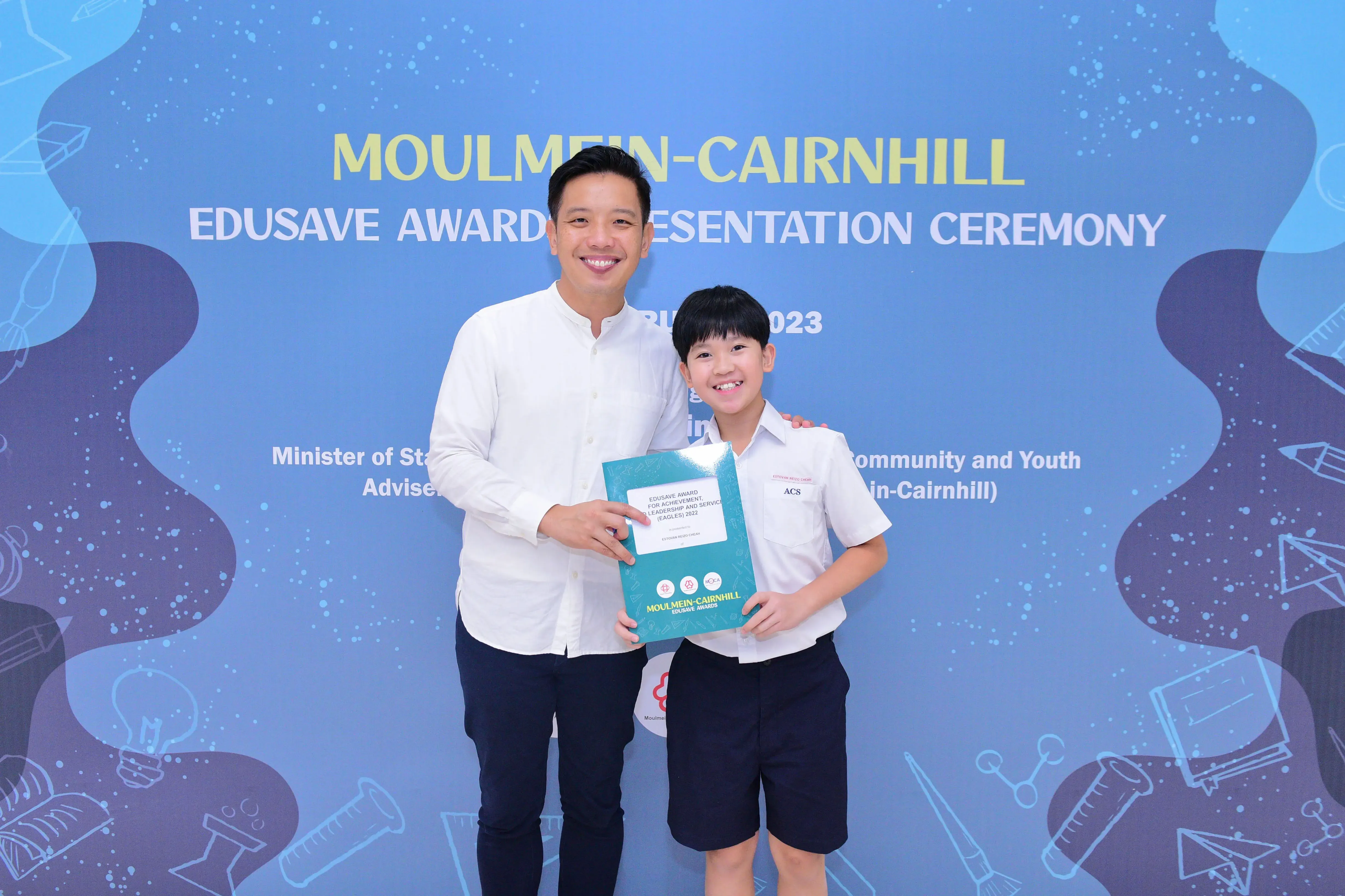 A man and a young boy in school uniform smile holding an award folder at the Moulmein-Cairnhill Edusave ceremony.