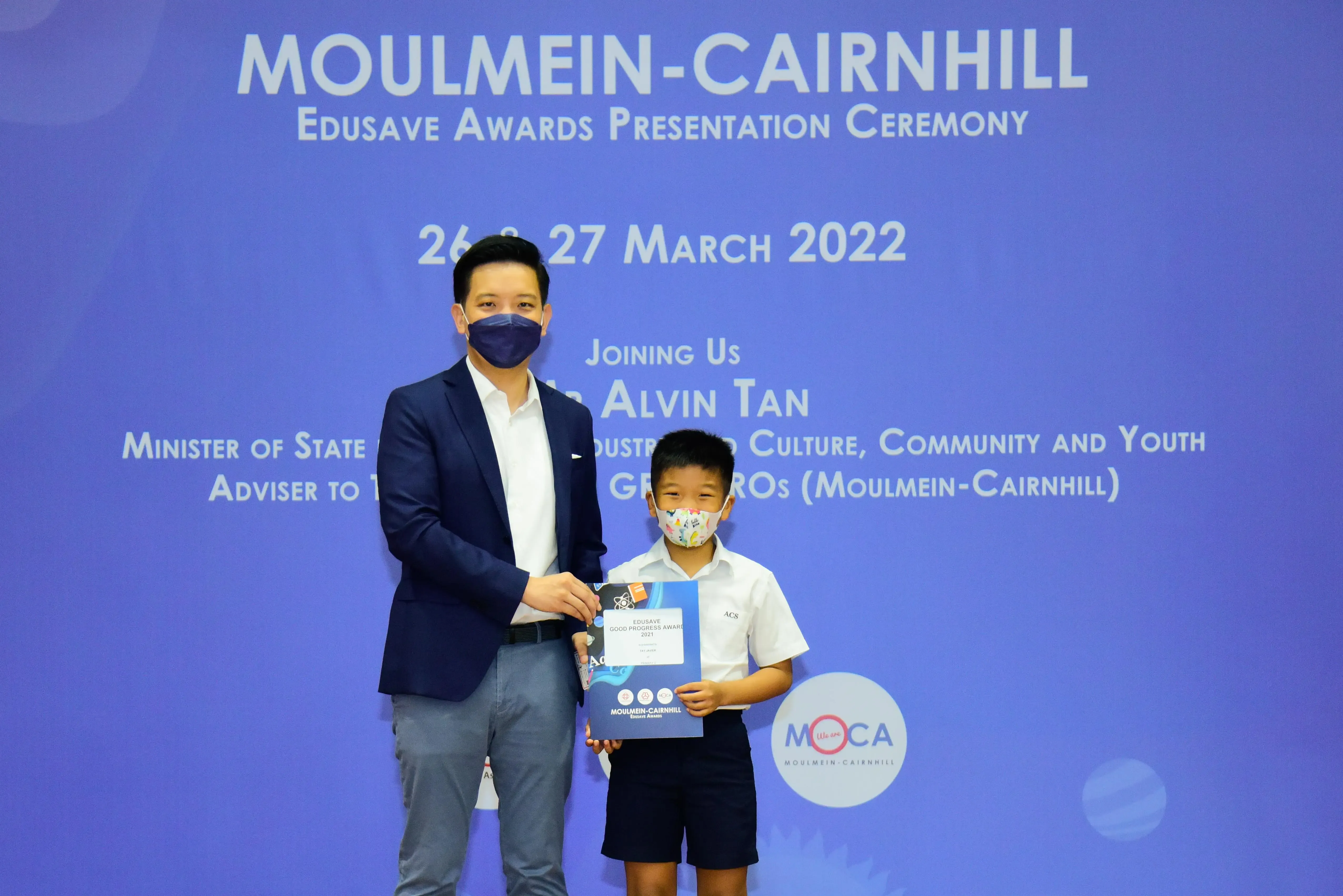An adult and a young boy wearing masks hold an Edusave award at the Moulmein-Cairnhill Awards Ceremony, March 2022.