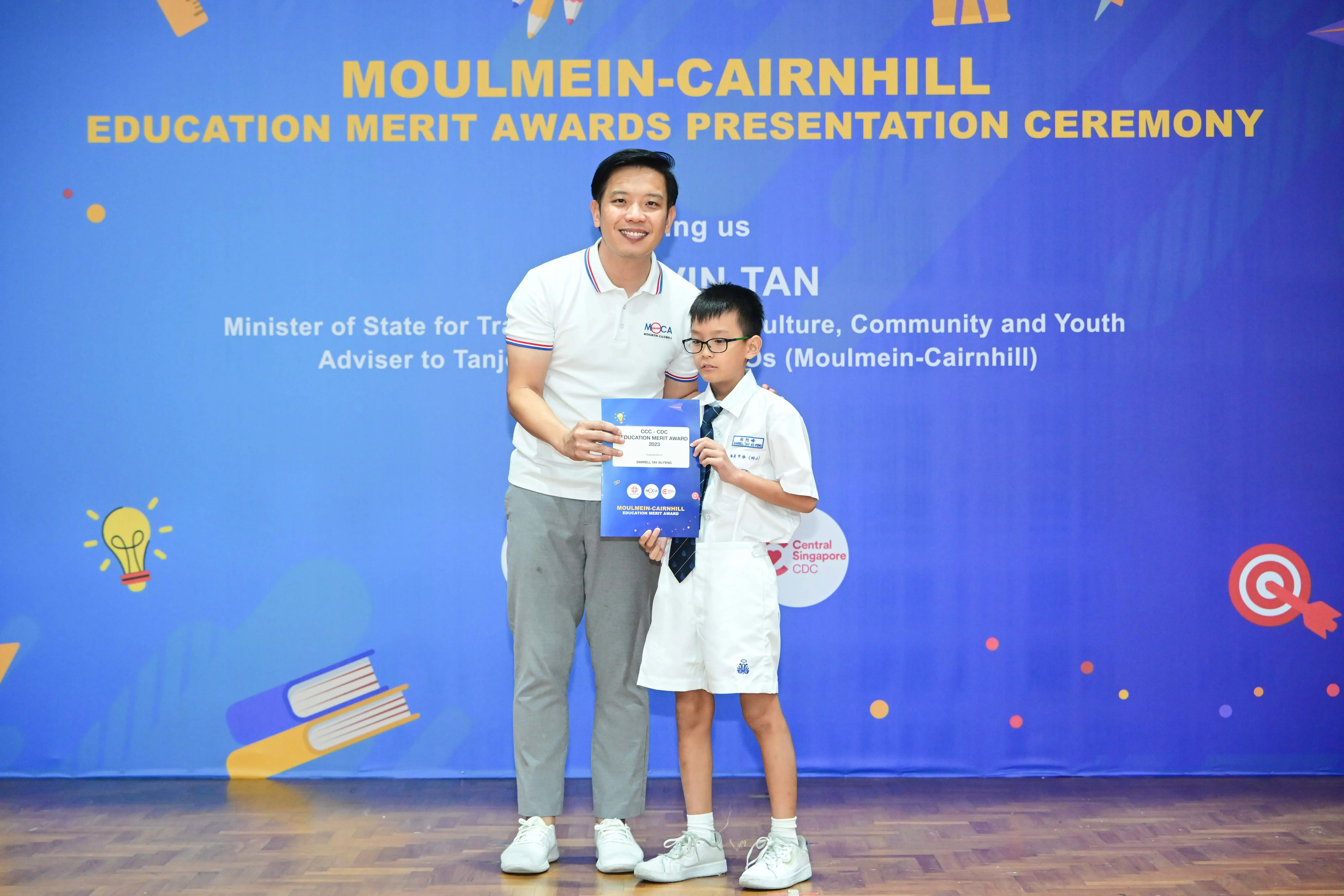 A man and a young boy in school uniform hold certificates at the Moulmein-Cairnhill Education Merit Awards ceremony.