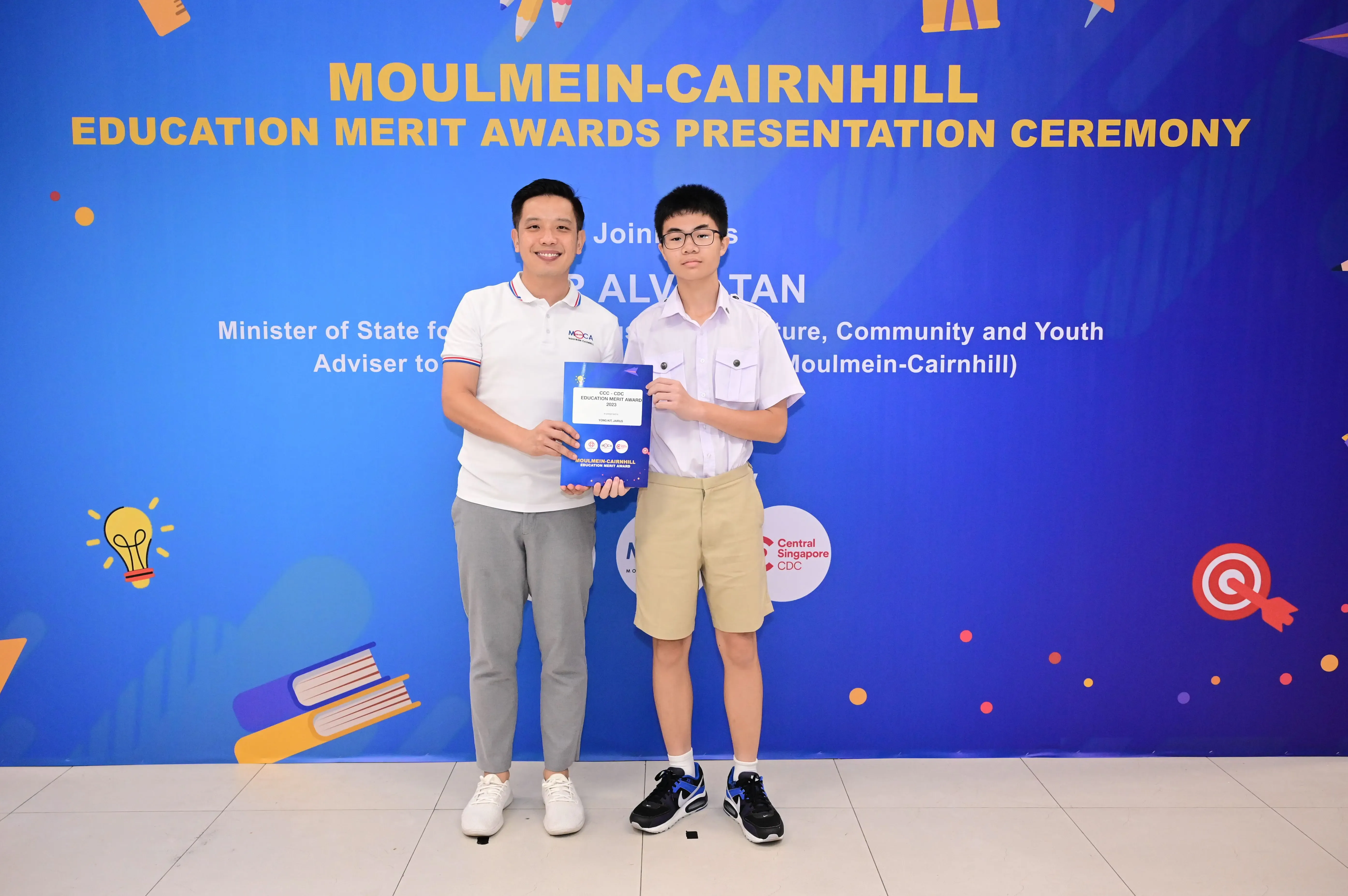 Two people hold an award certificate in front of a blue Moulmein-Cairnhill Education Merit Awards banner.