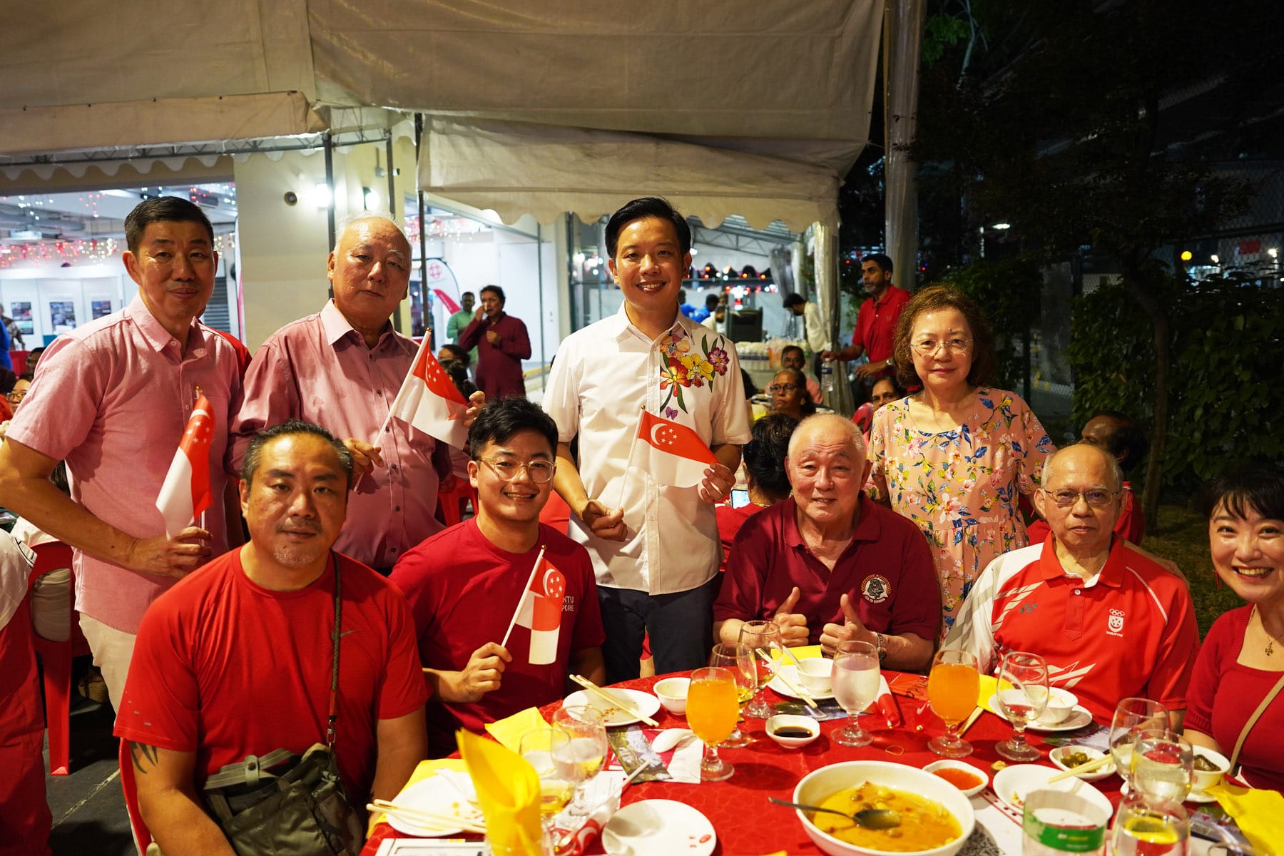 Group of people in red and white clothing posing at an outdoor dinner table, holding small Singapore flags at night.