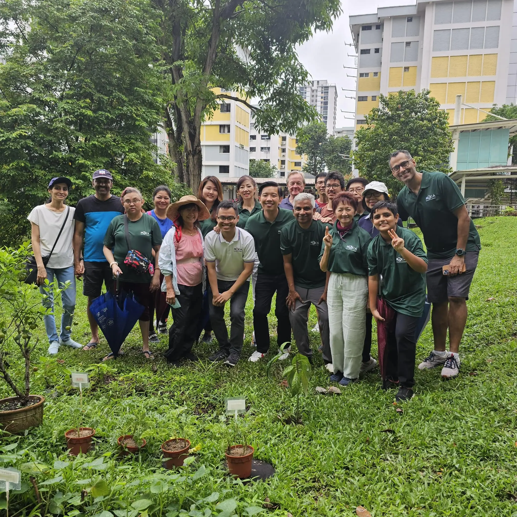 A group of about 15 people posing outdoors on grass near potted plants, trees, and colorful urban buildings.