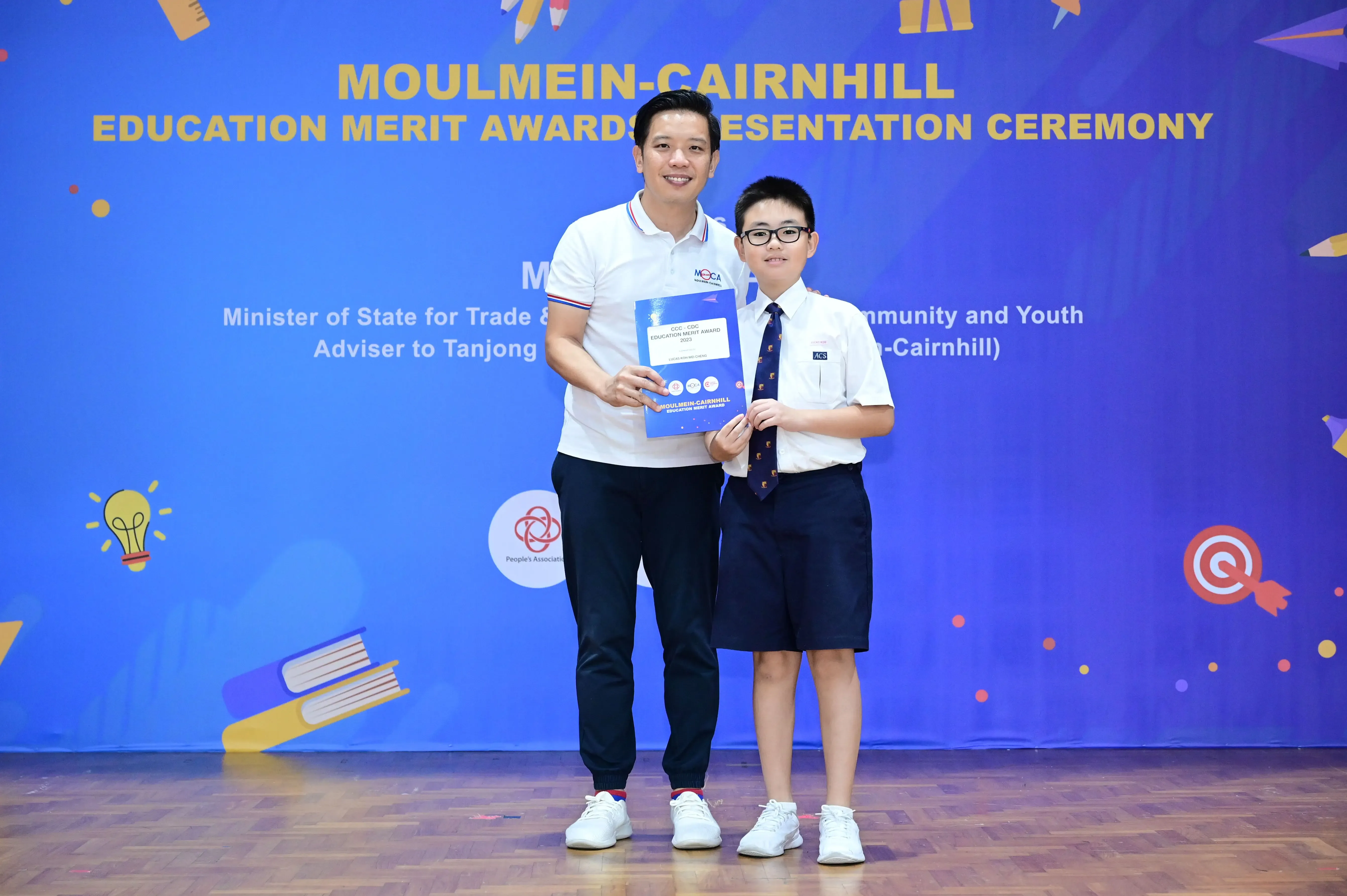 An adult and a young student in school uniform hold an award certificate at the Moulmein-Cairnhill Education Merit Awards ceremony.