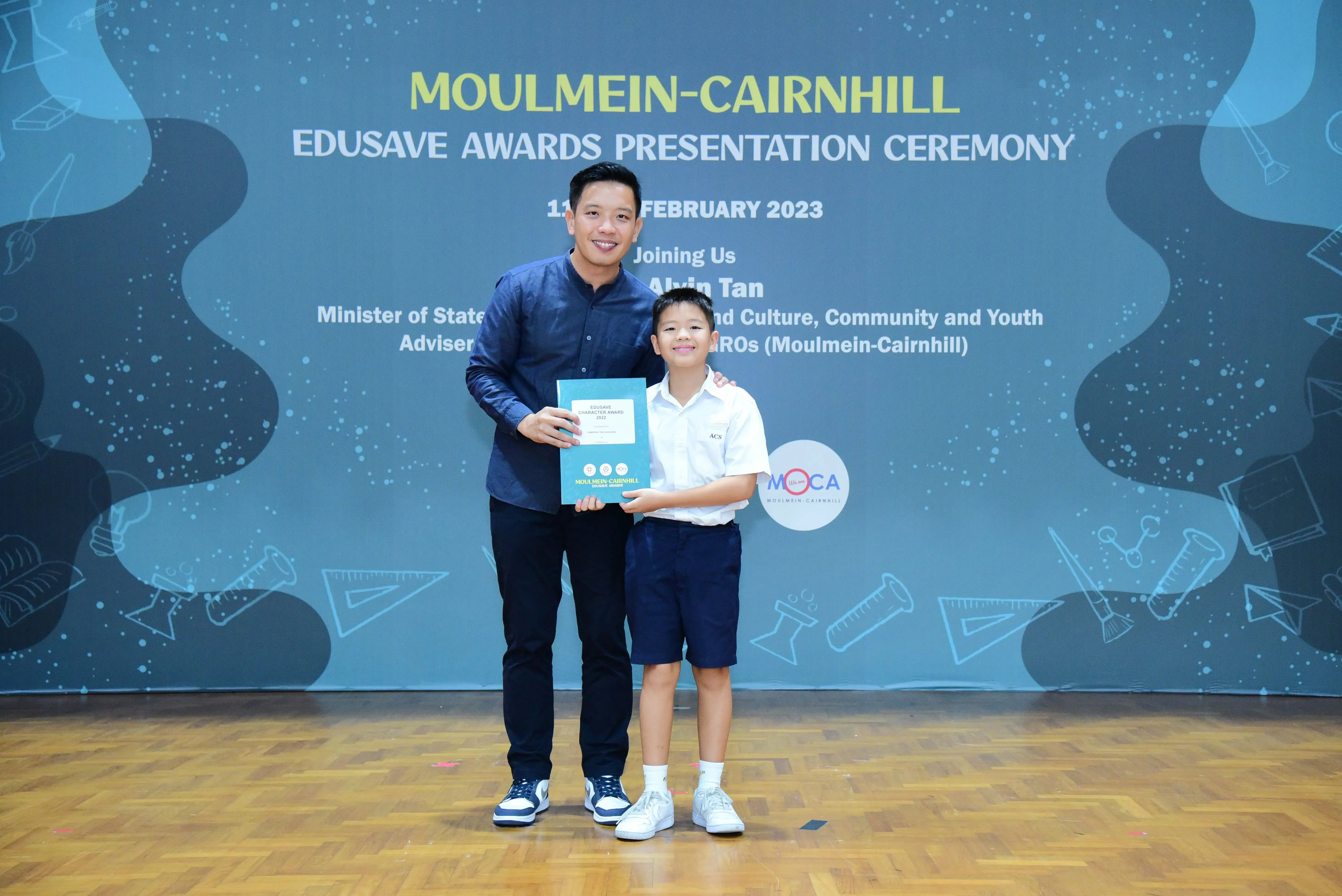 An adult and a young boy hold a certificate at the Moulmein-Cairnhill Edusave Awards Presentation Ceremony 2023.