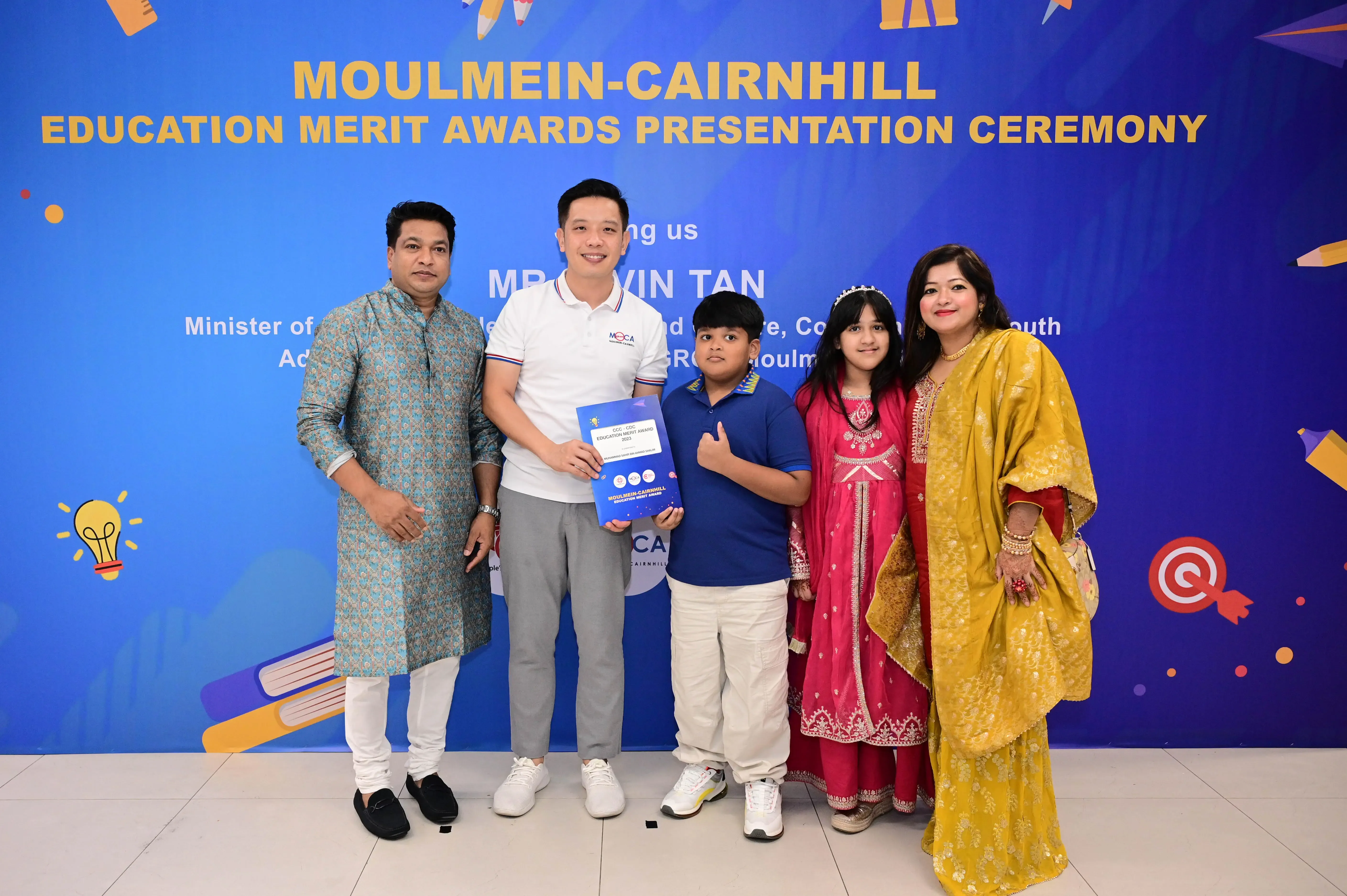 Five people pose at the Moulmein-Cairnhill Education Merit Awards Presentation Ceremony, with a boy holding an award certificate.