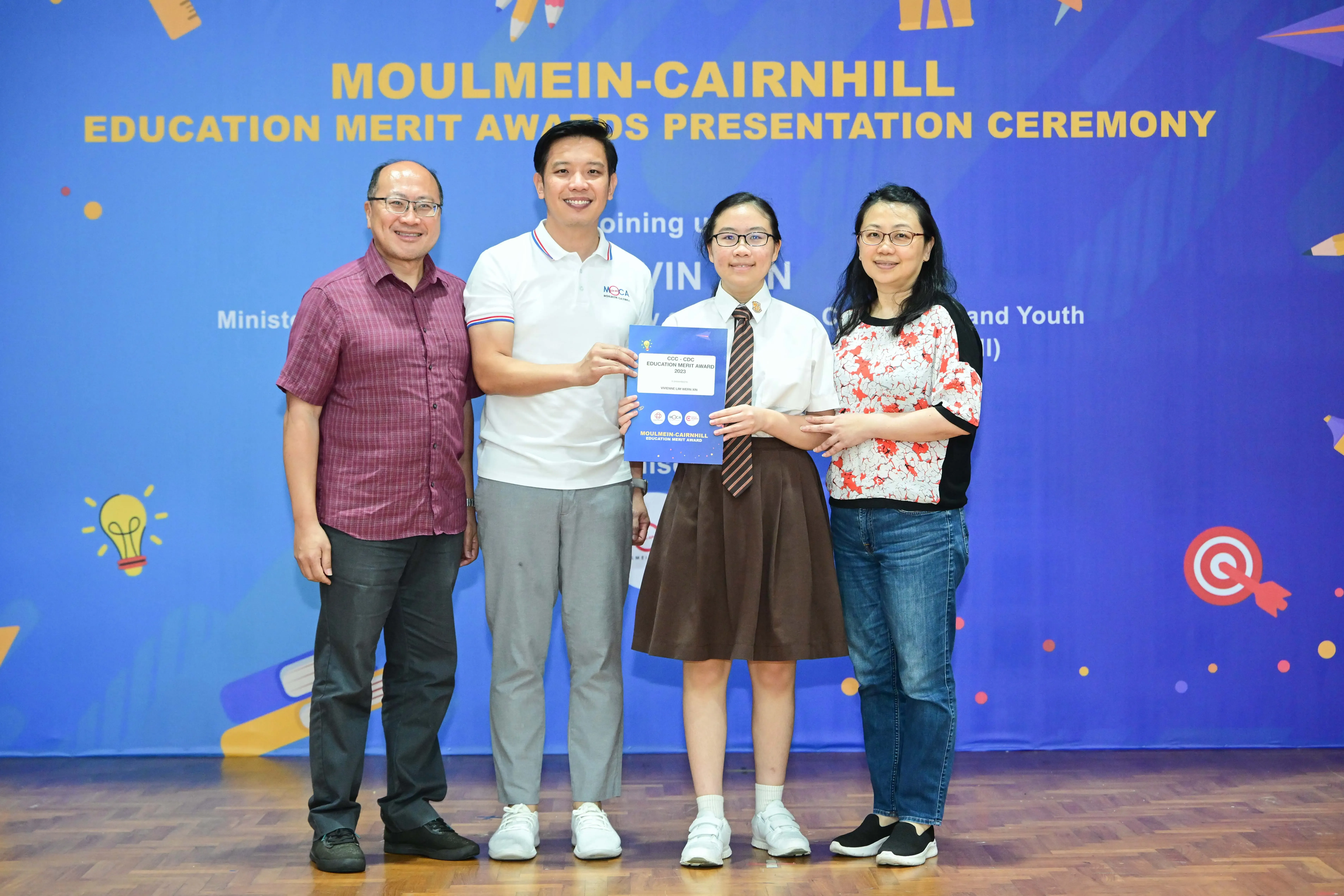 Four people pose at the Moulmein-Cairnhill Education Merit Awards Presentation Ceremony, with a student holding a certificate.