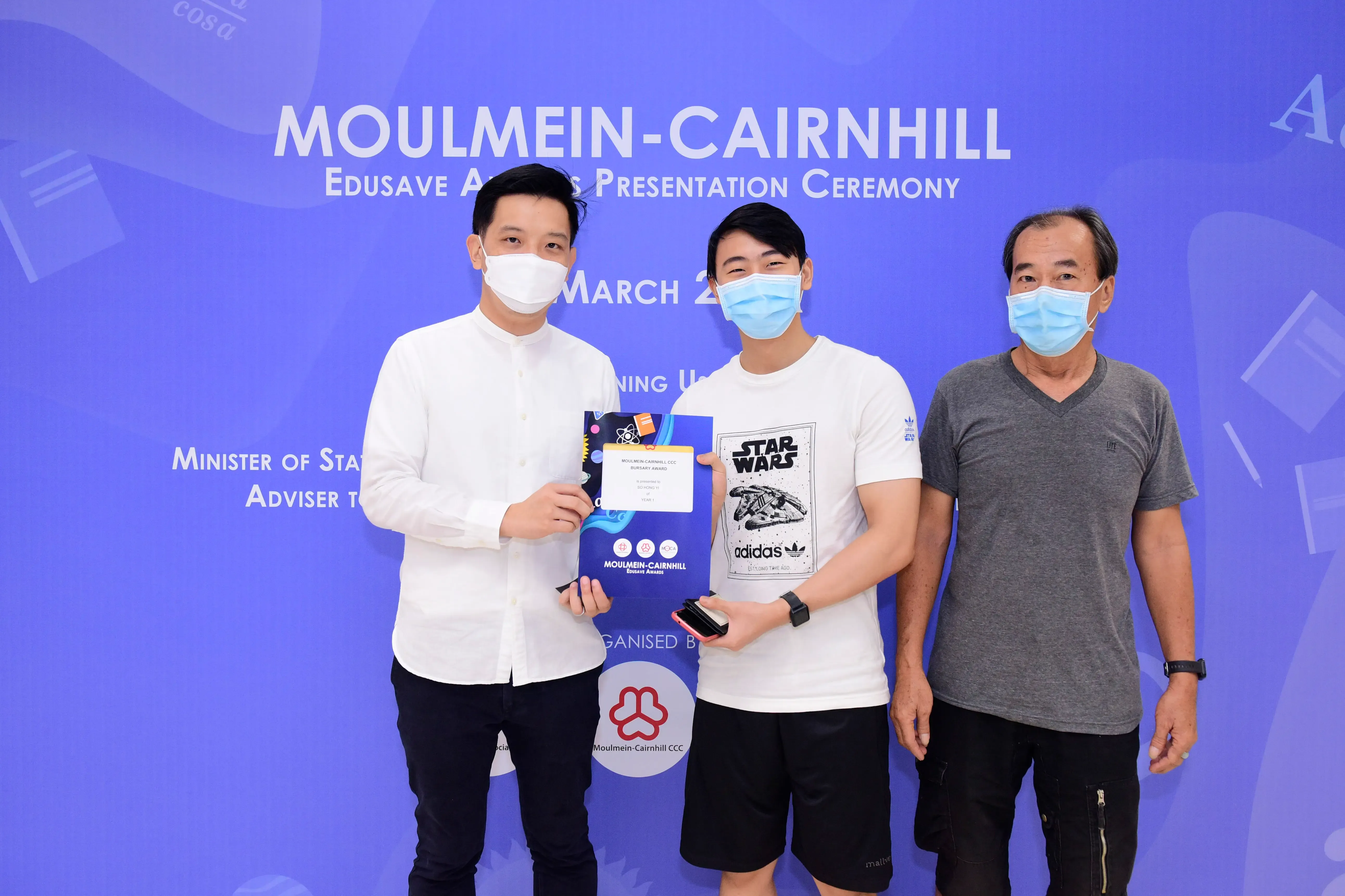 Three masked men pose at the Moulmein-Cairnhill Edusave Awards Presentation Ceremony, one holding a certificate.