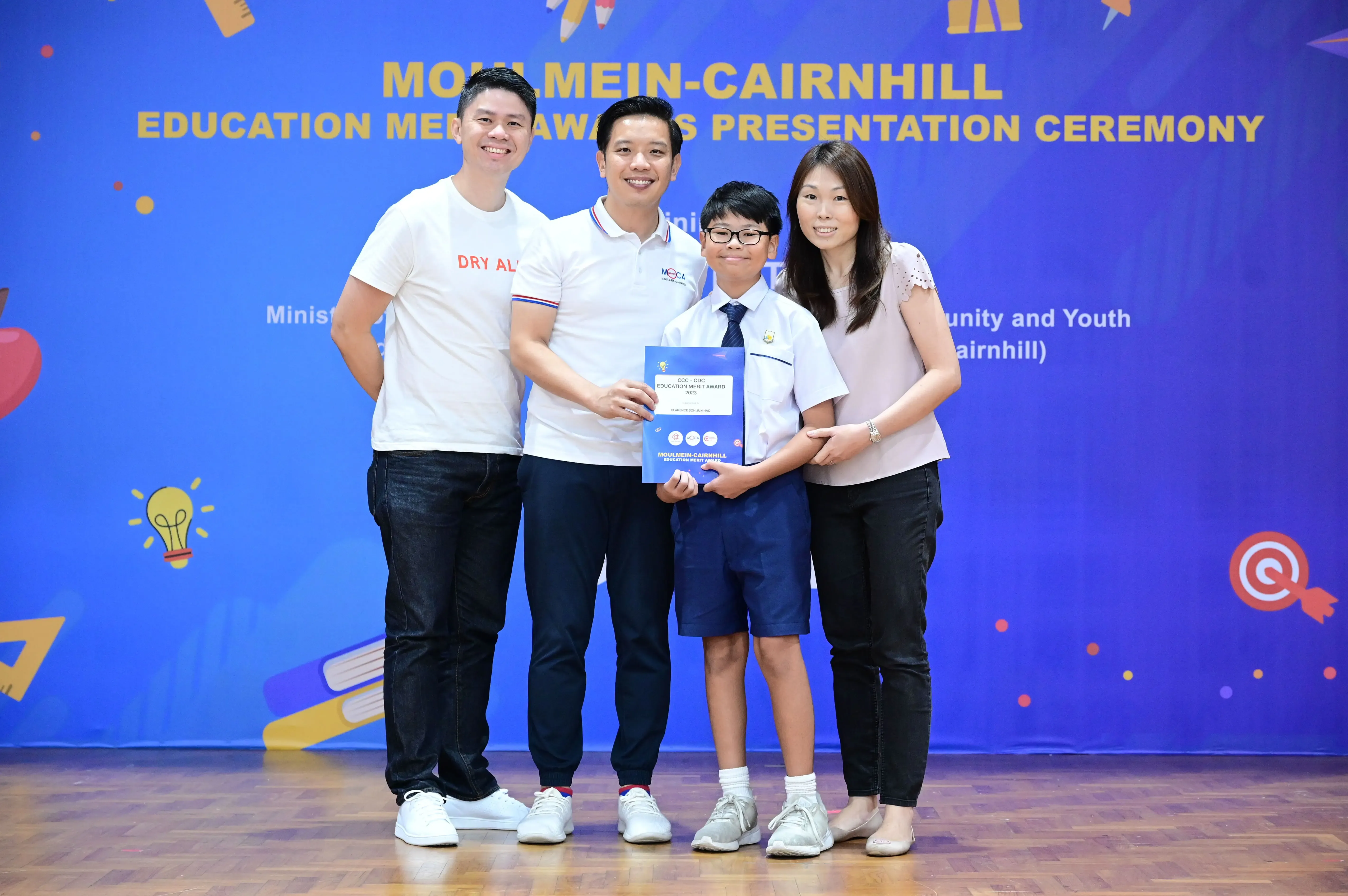 Three adults and a boy in school uniform hold an award certificate at the Moulmein-Cairnhill Education Merit Awards ceremony.