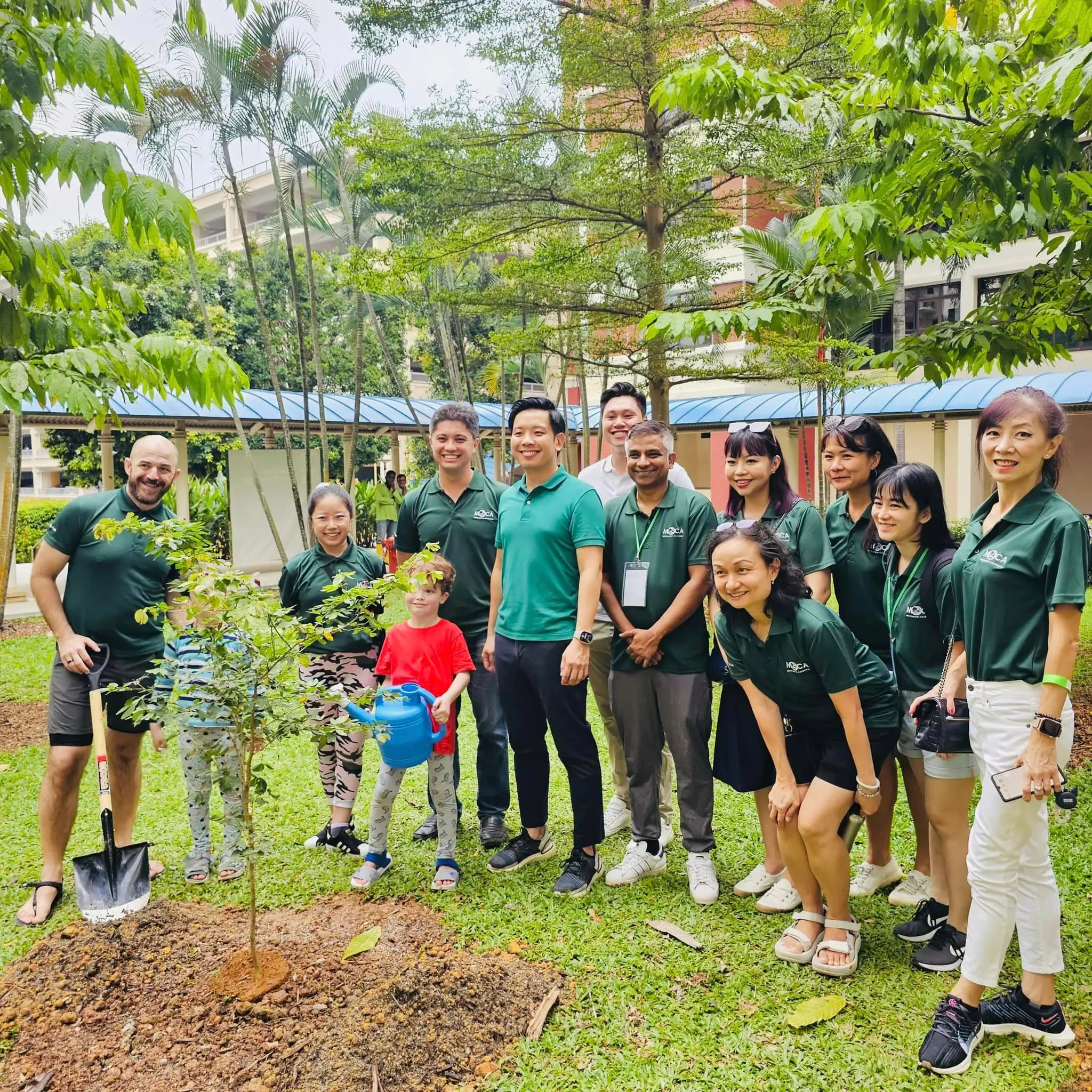 A group of adults and children in green polo shirts pose outdoors near a freshly planted sapling with a shovel and watering can.