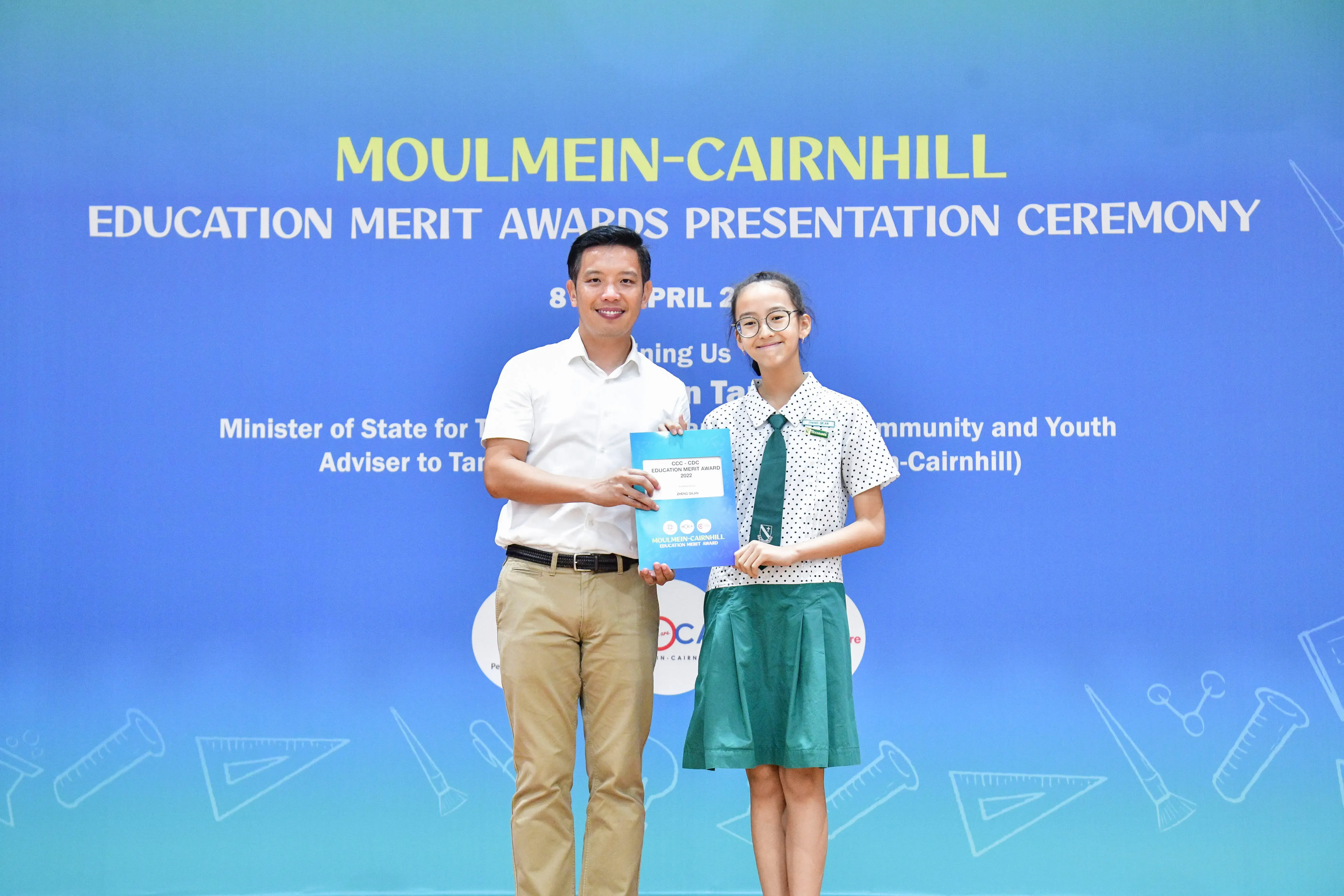 A man and a young girl in school uniform hold a blue certificate in front of a Moulmein-Cairnhill awards banner.