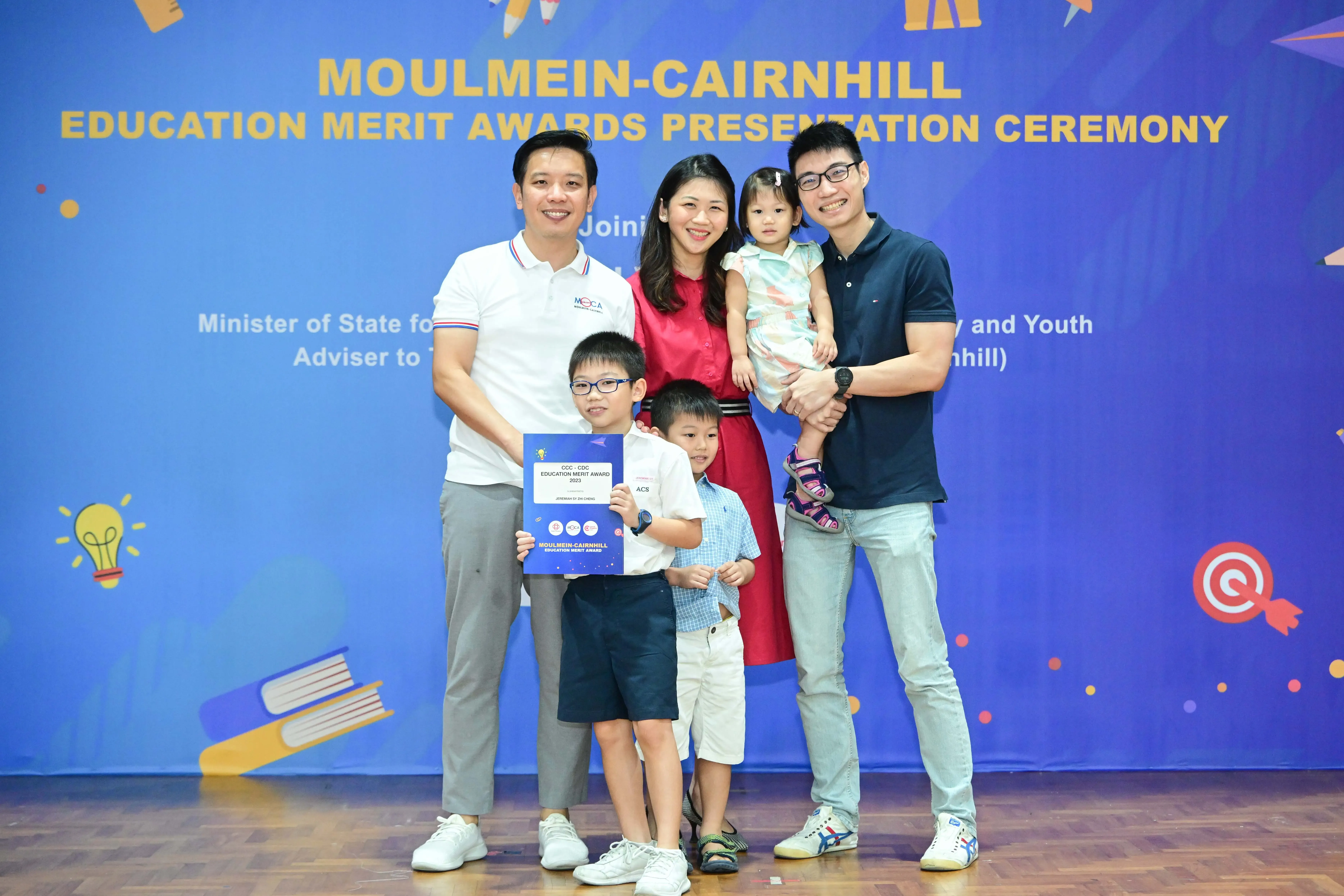 Family of five posing at the Moulmein-Cairnhill Education Merit Awards Presentation Ceremony, child holding a blue award certificate.