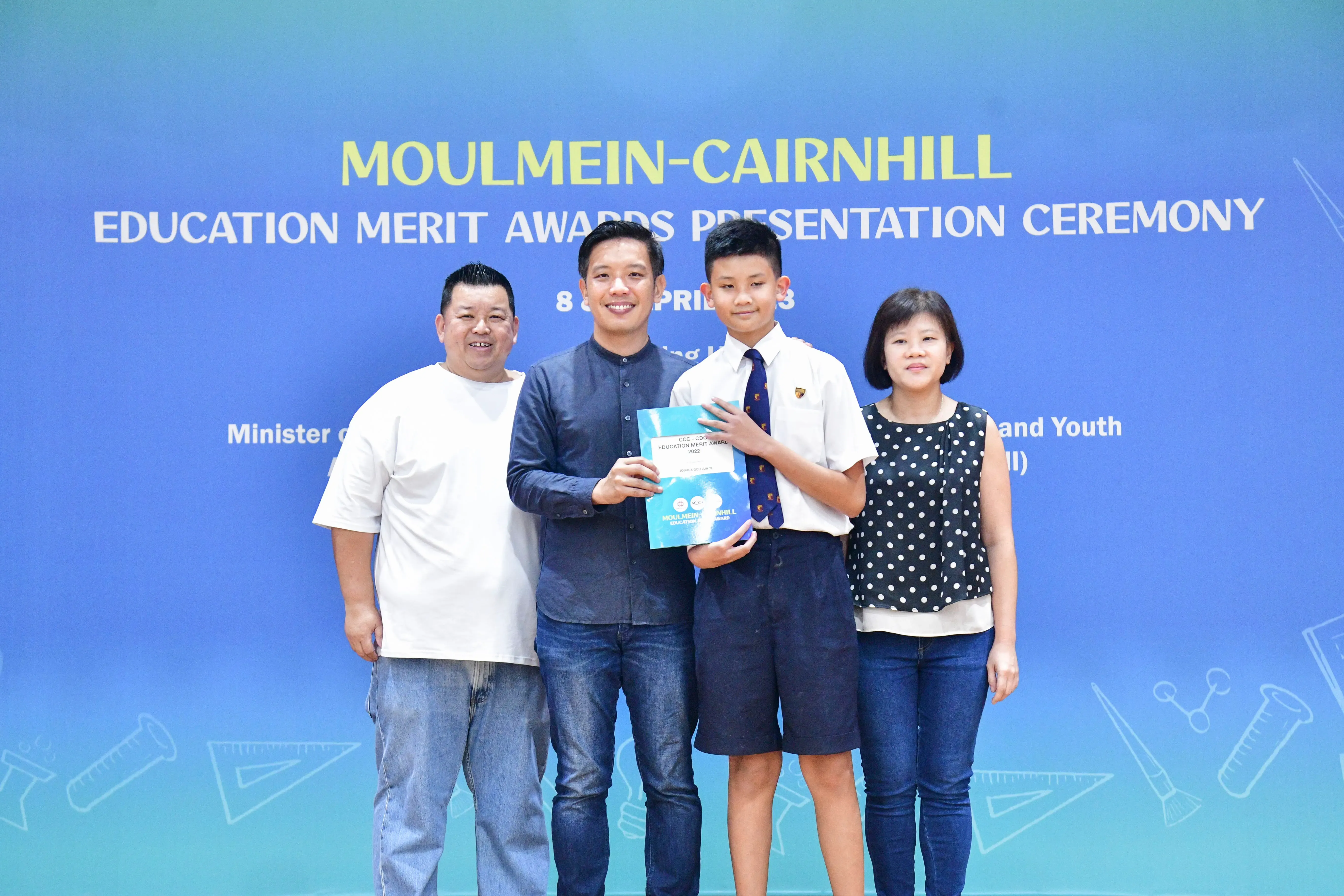 Four people pose at the Moulmein-Cairnhill Education Merit Awards Presentation Ceremony, holding a blue award booklet.