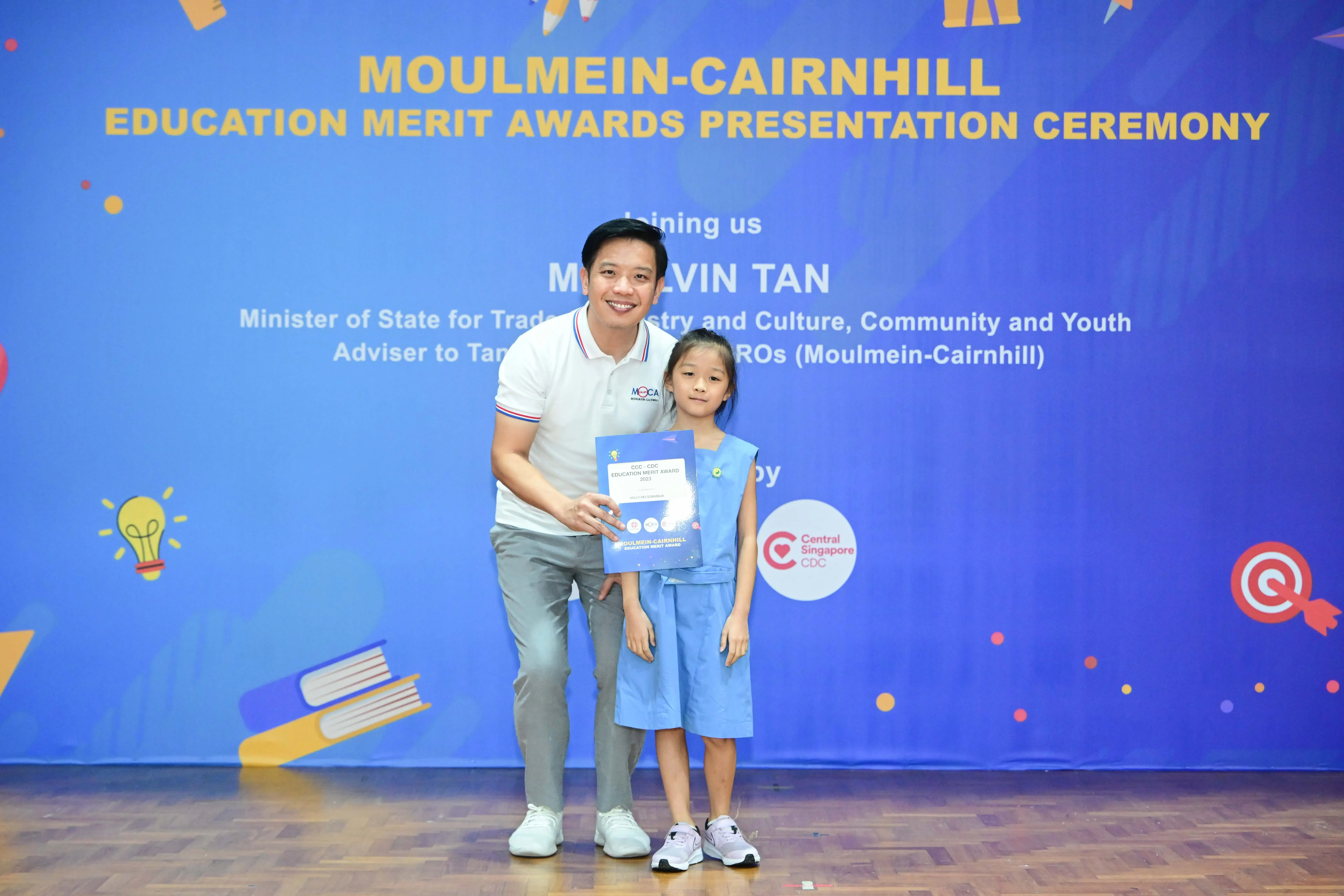 A man and a young girl hold a certificate at the Moulmein-Cairnhill Education Merit Awards Presentation Ceremony.