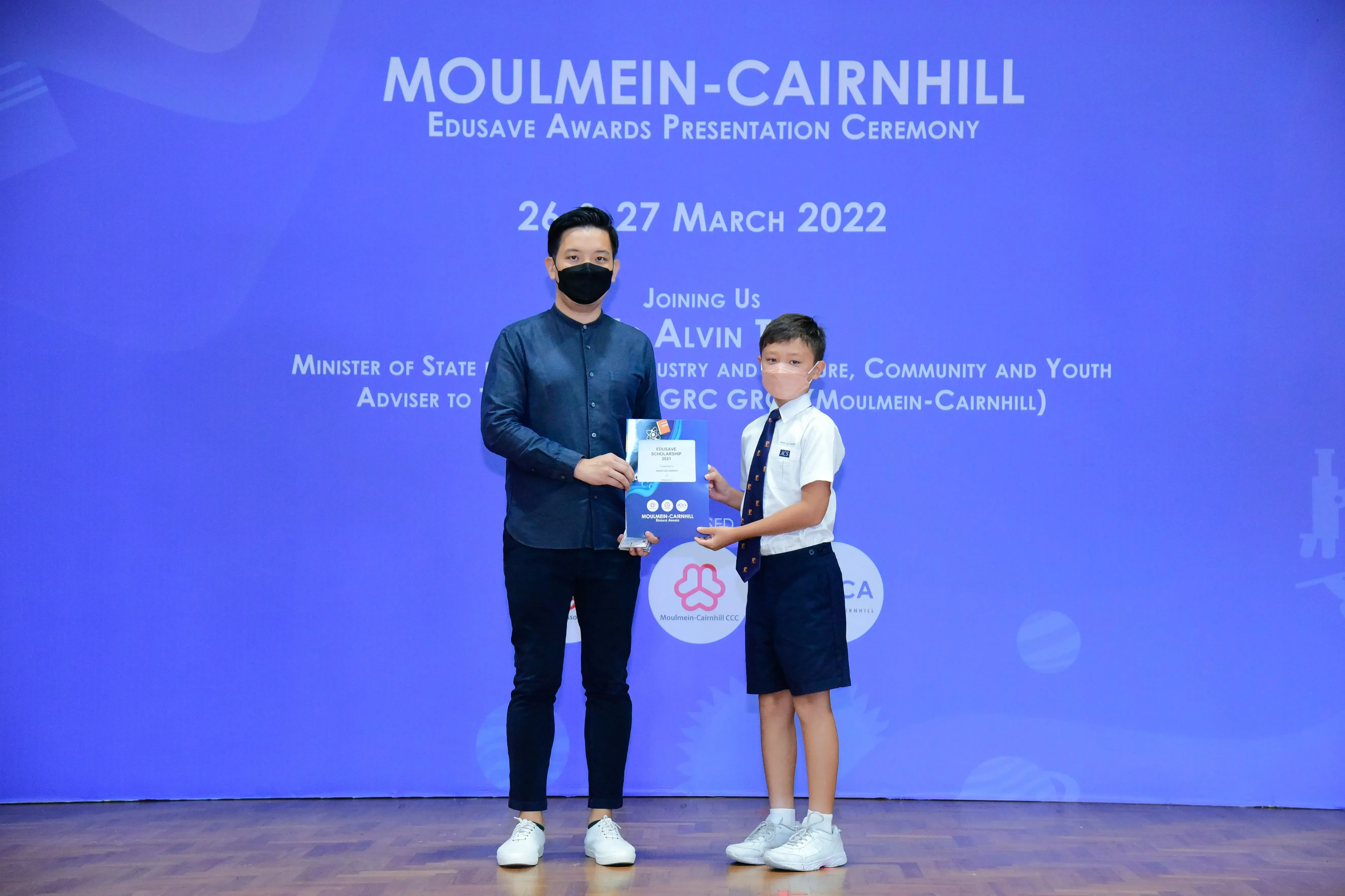 An adult and a young boy in school uniform hold an award at the Moulmein-Cairnhill Edusave Awards Ceremony 2022.