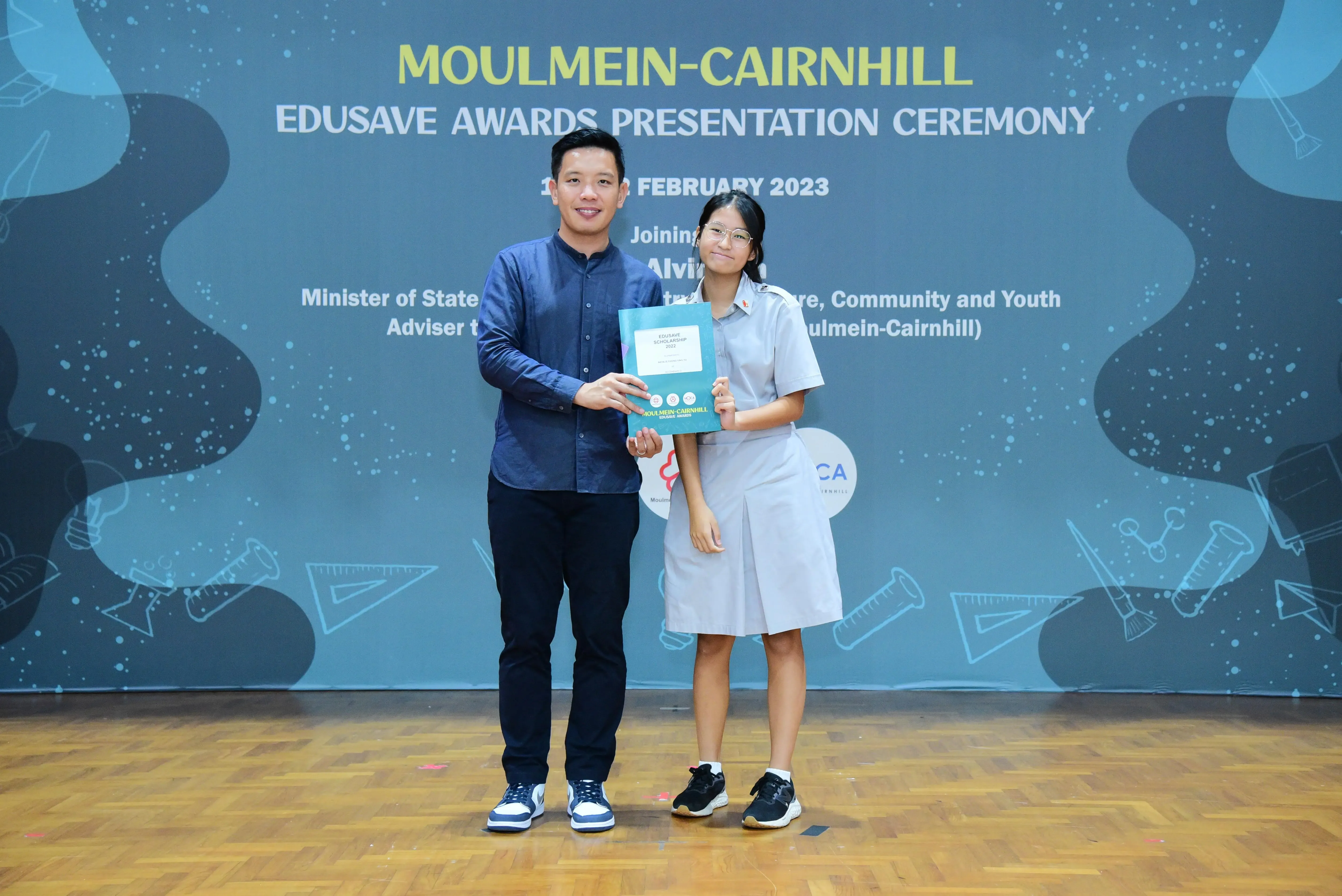 A man and a student in school uniform hold a certificate at the Moulmein-Cairnhill Edusave Awards Ceremony, February 2023.