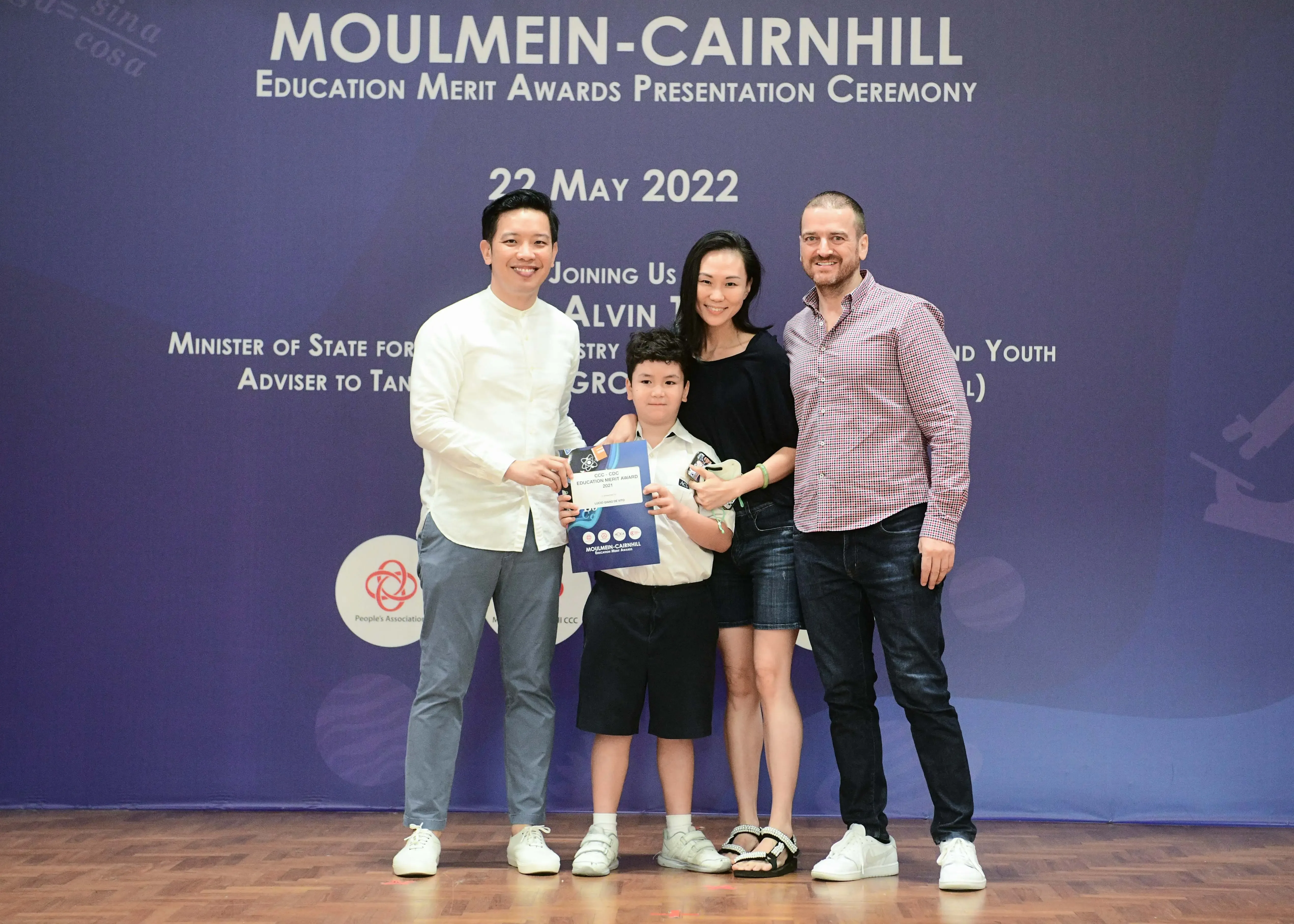 Four people pose before a Moulmein-Cairnhill Education Merit Awards banner dated 22 May 2022, a boy holds a certificate.