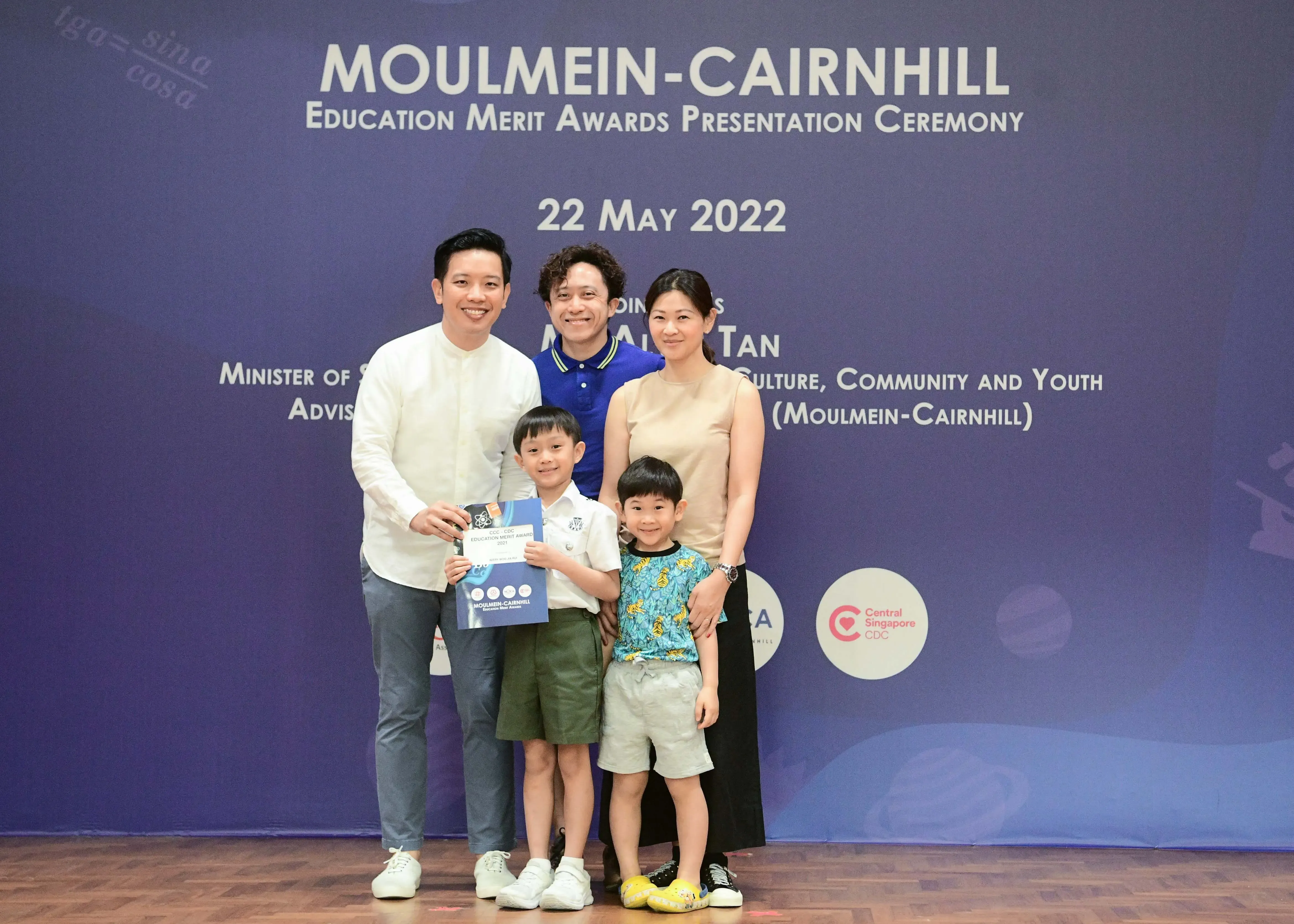 Five people pose before a Moulmein-Cairnhill Education Merit Awards banner dated 22 May 2022.