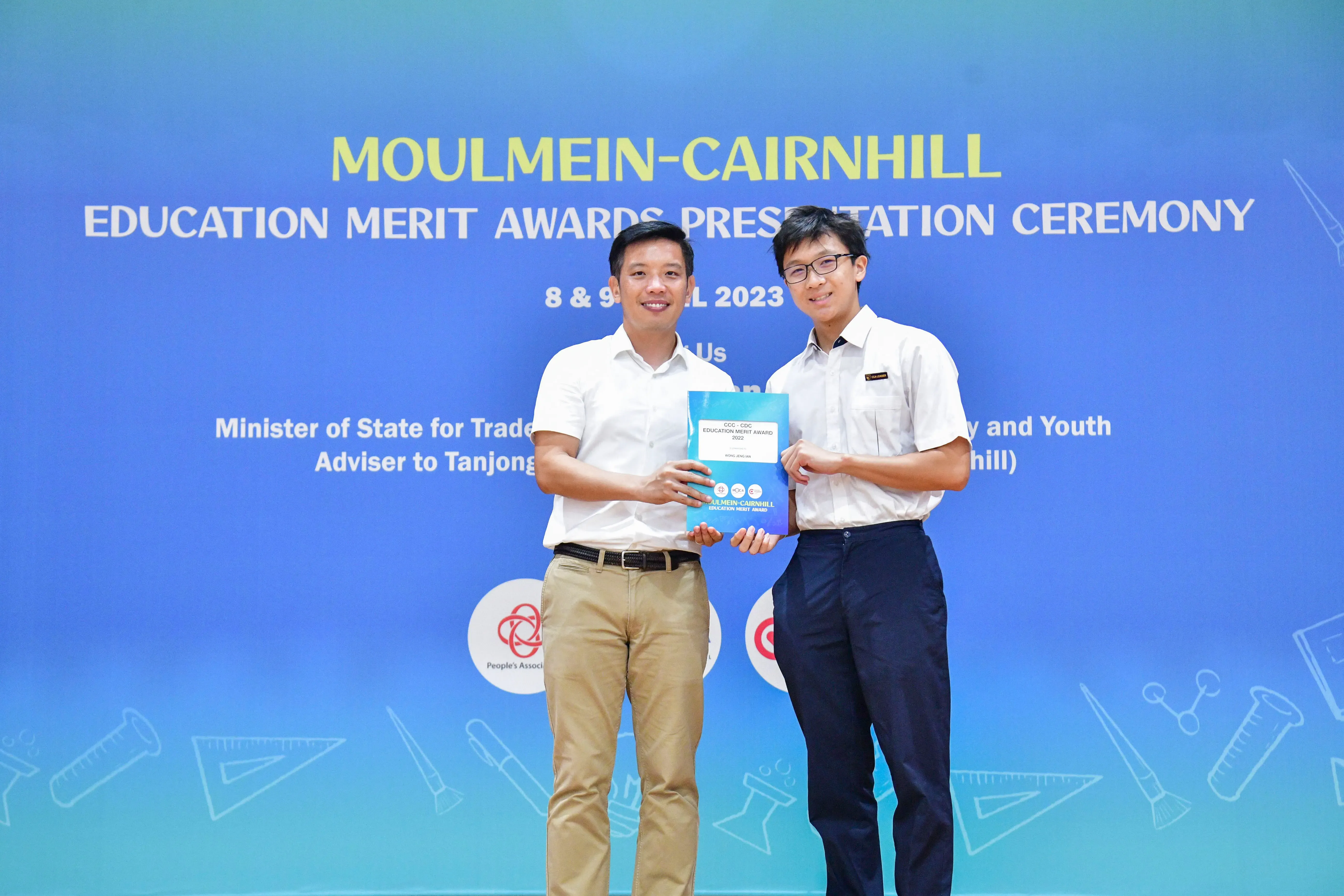 Two men hold a certificate together in front of a Moulmein-Cairnhill Education Merit Awards 2023 banner.