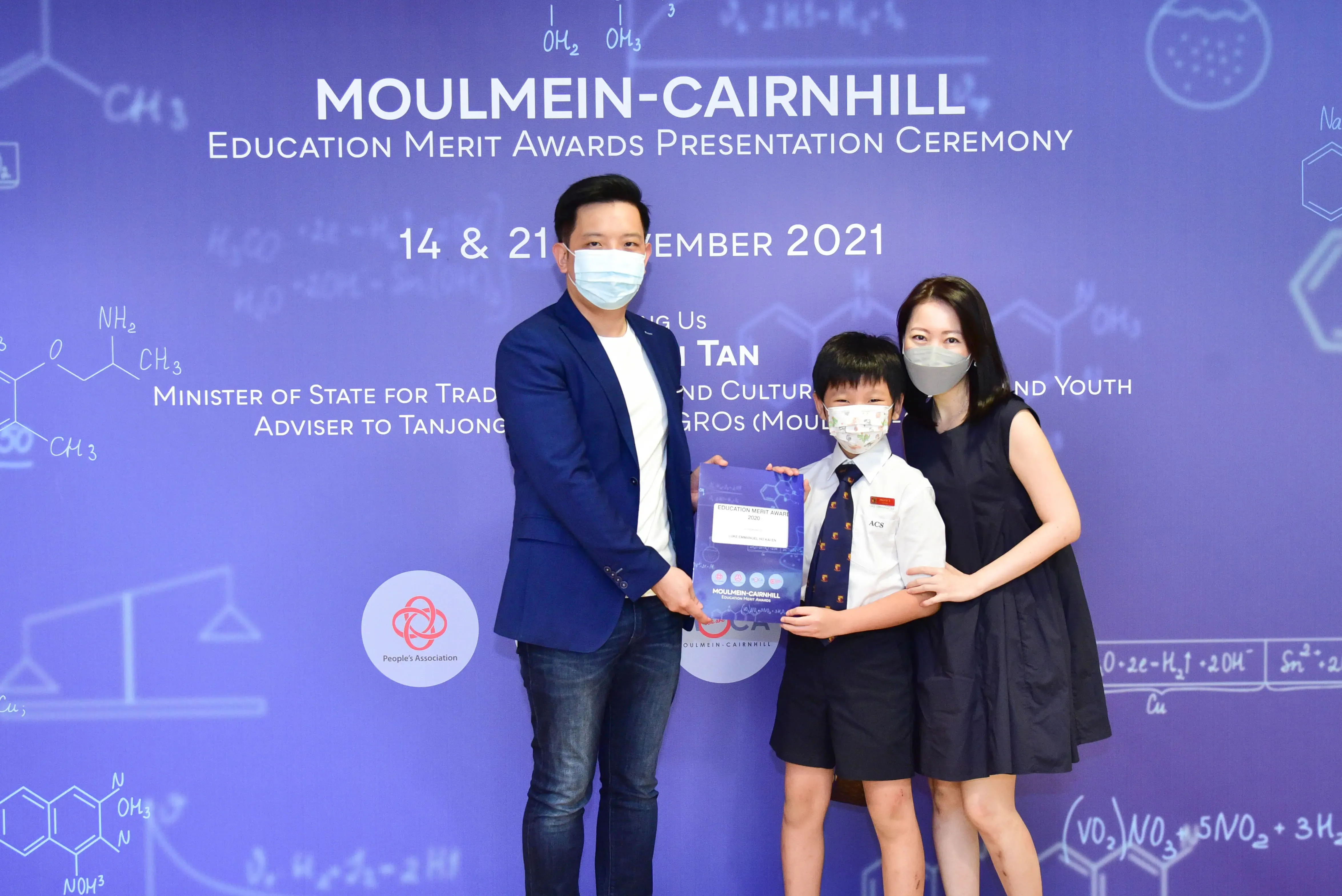 Two masked adults and a masked child in school uniform hold a certificate at the Moulmein-Cairnhill Education Merit Awards 2021.