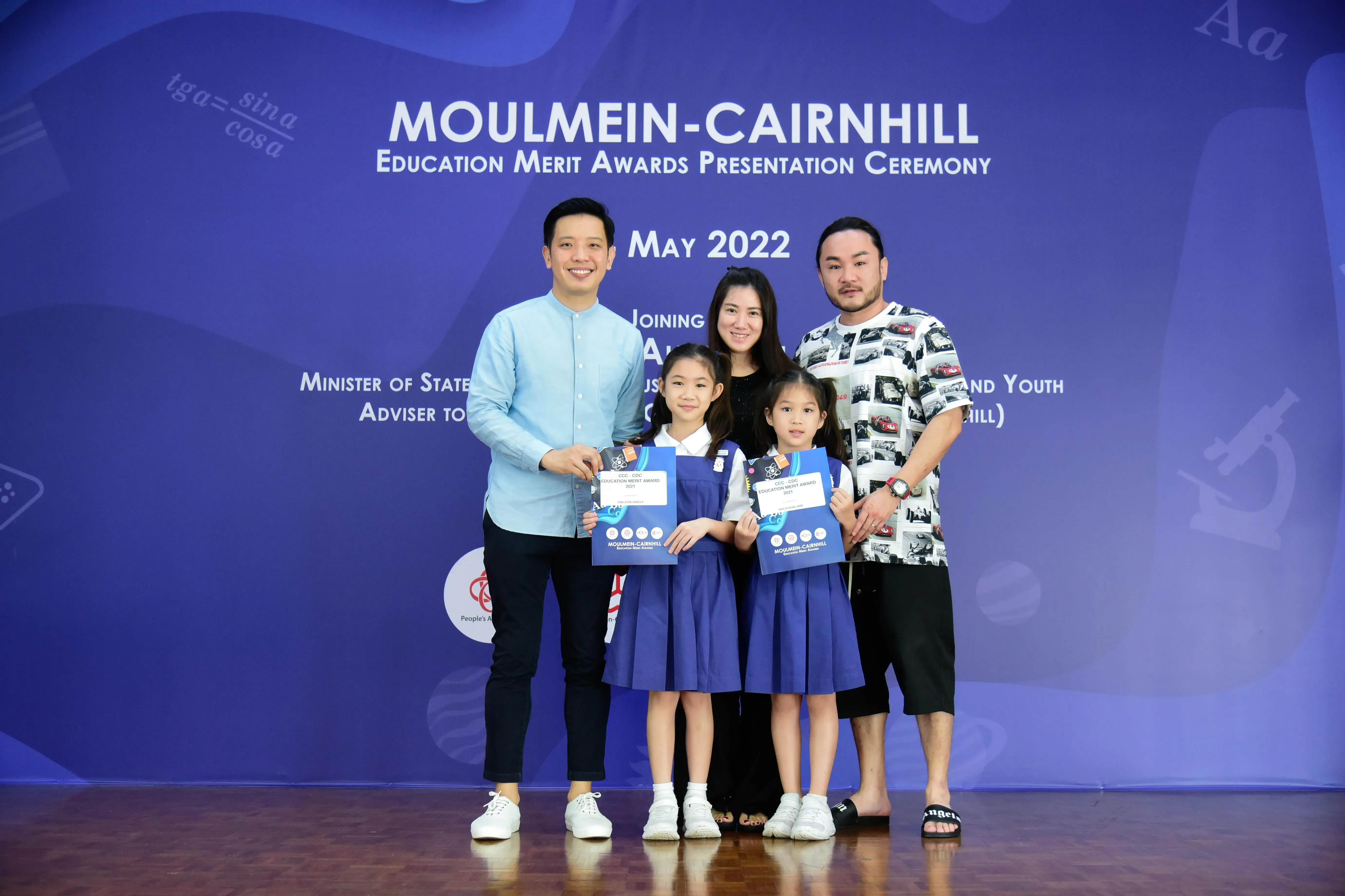 Two adults and two girls in blue uniforms hold award folders at the Moulmein-Cairnhill Education Merit Awards May 2022.