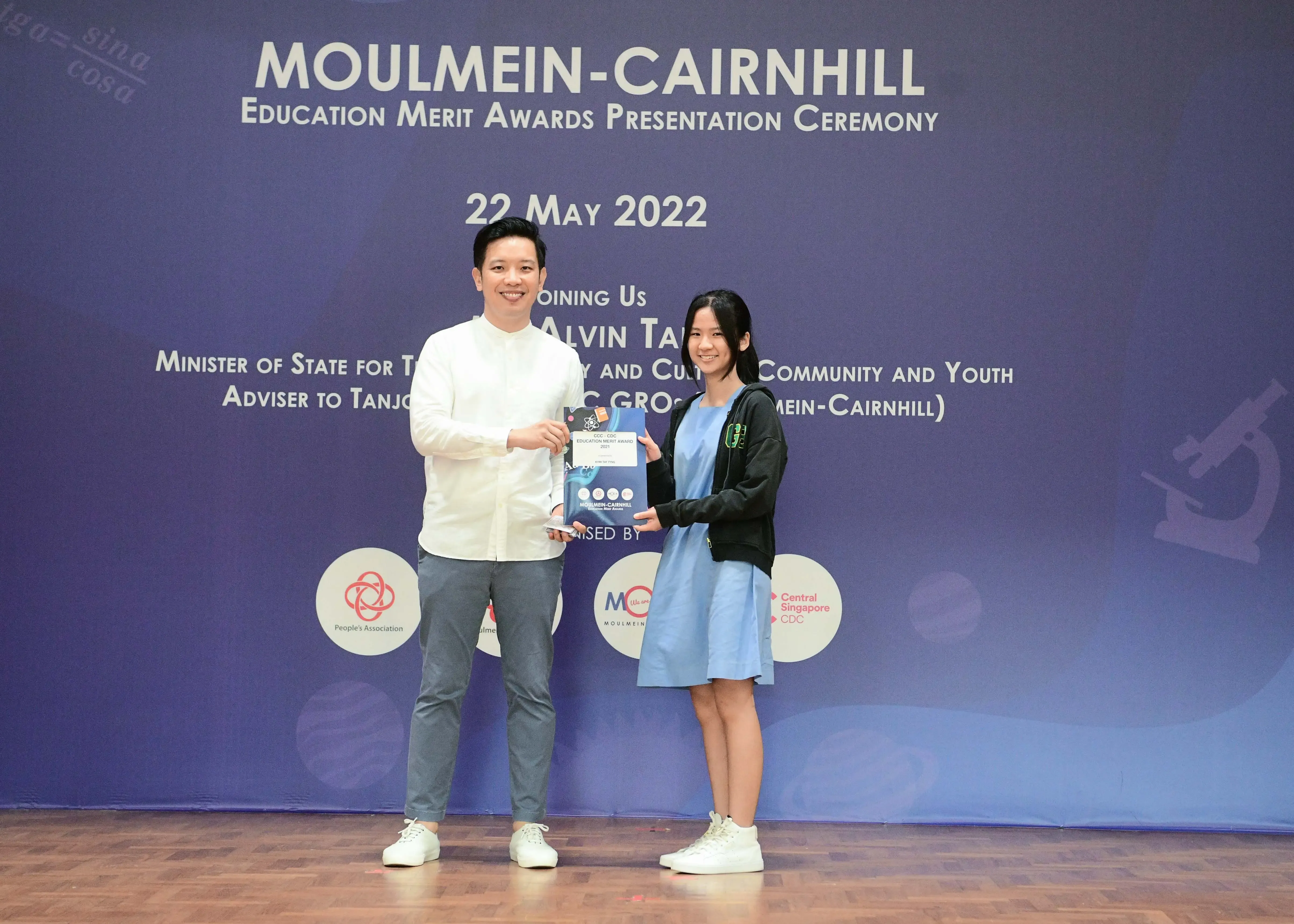 Two people hold an award together on stage at the Moulmein-Cairnhill Education Merit Awards ceremony on 22 May 2022.