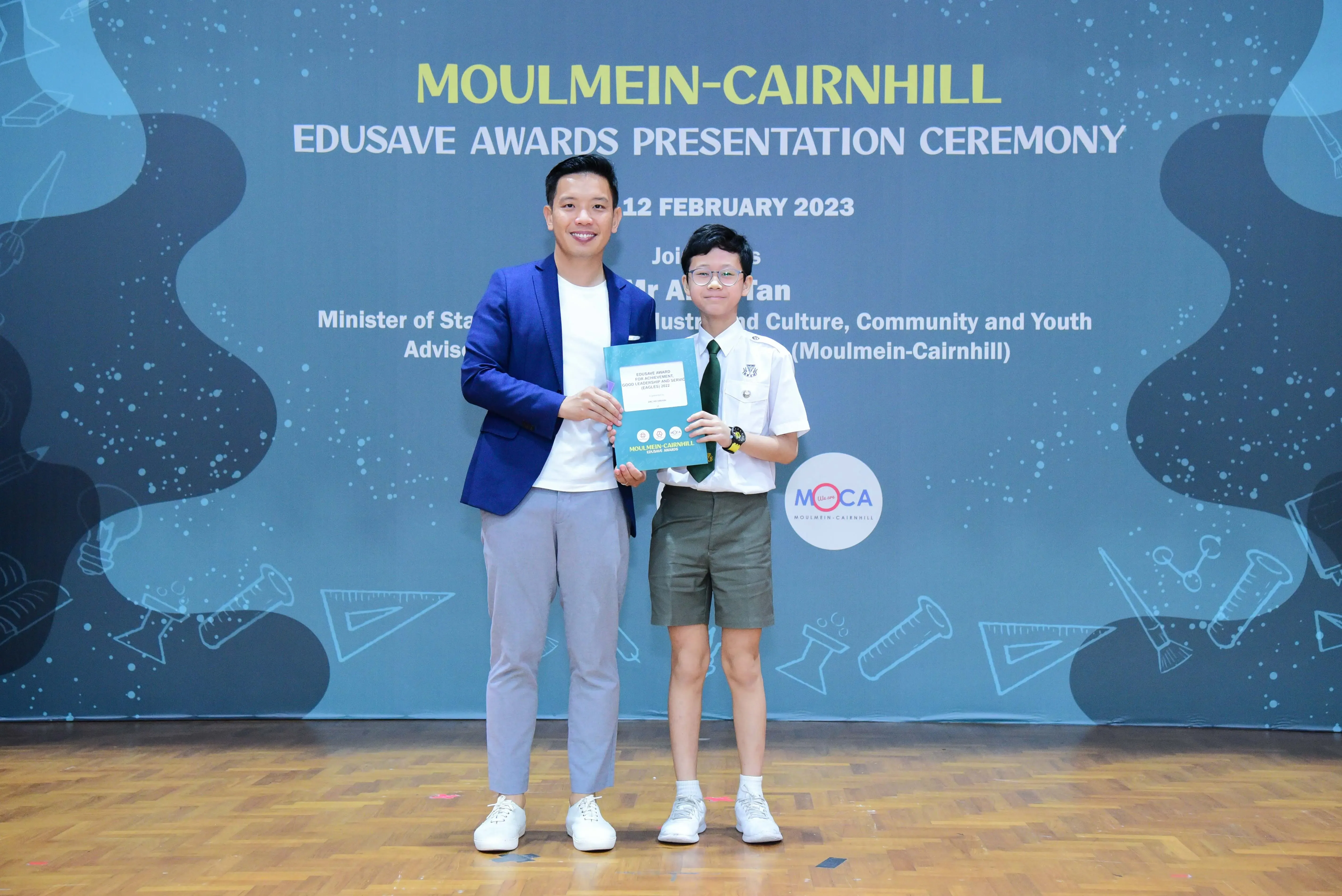 An adult in a blue blazer and a uniformed student hold a certificate at the Moulmein-Cairnhill Edusave Awards 2023.