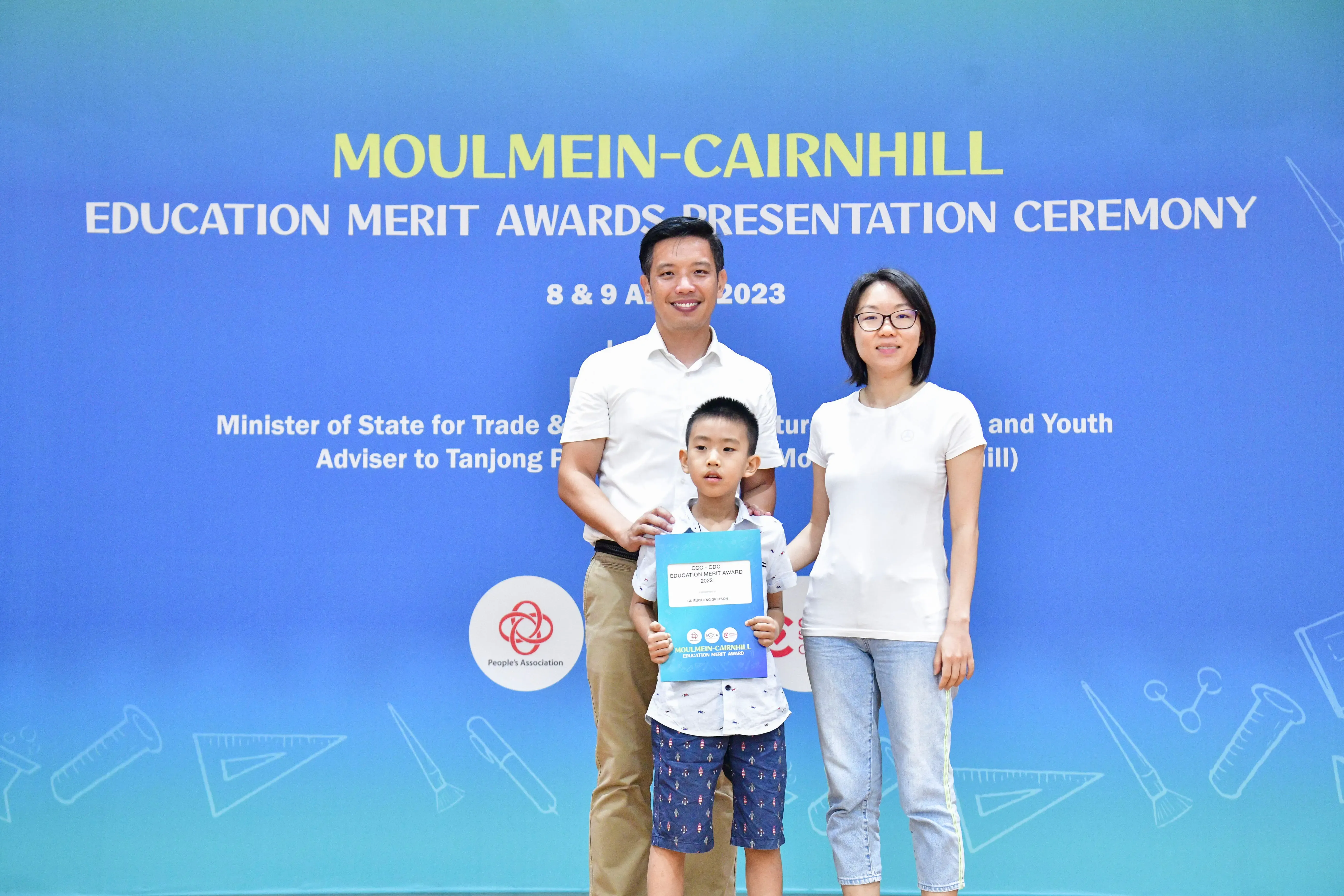 A man, woman, and young boy pose before a Moulmein-Cairnhill Education Merit Awards Ceremony banner; the boy holds a certificate.