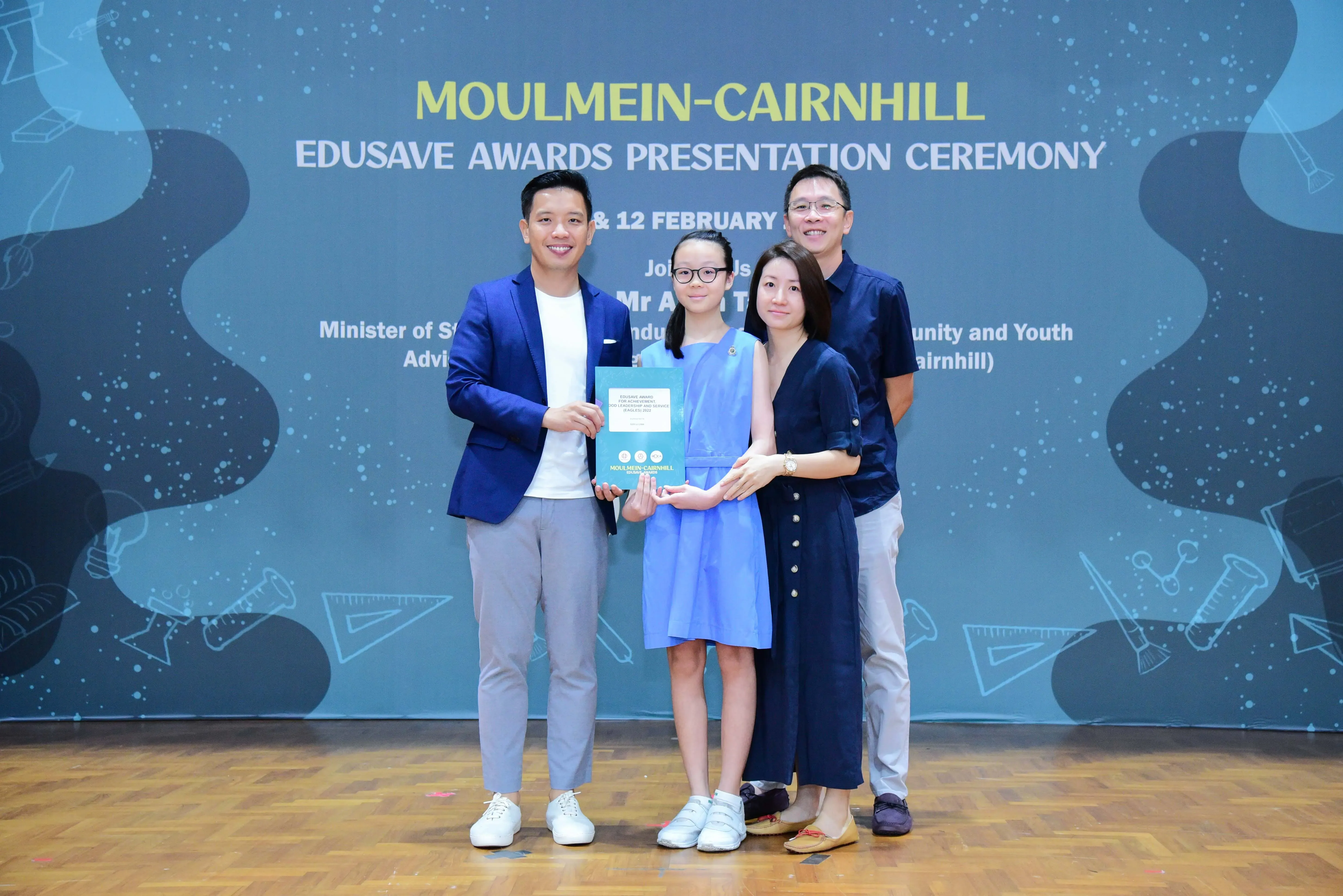 Four people pose holding a certificate at the Moulmein-Cairnhill Edusave Awards Presentation Ceremony backdrop.