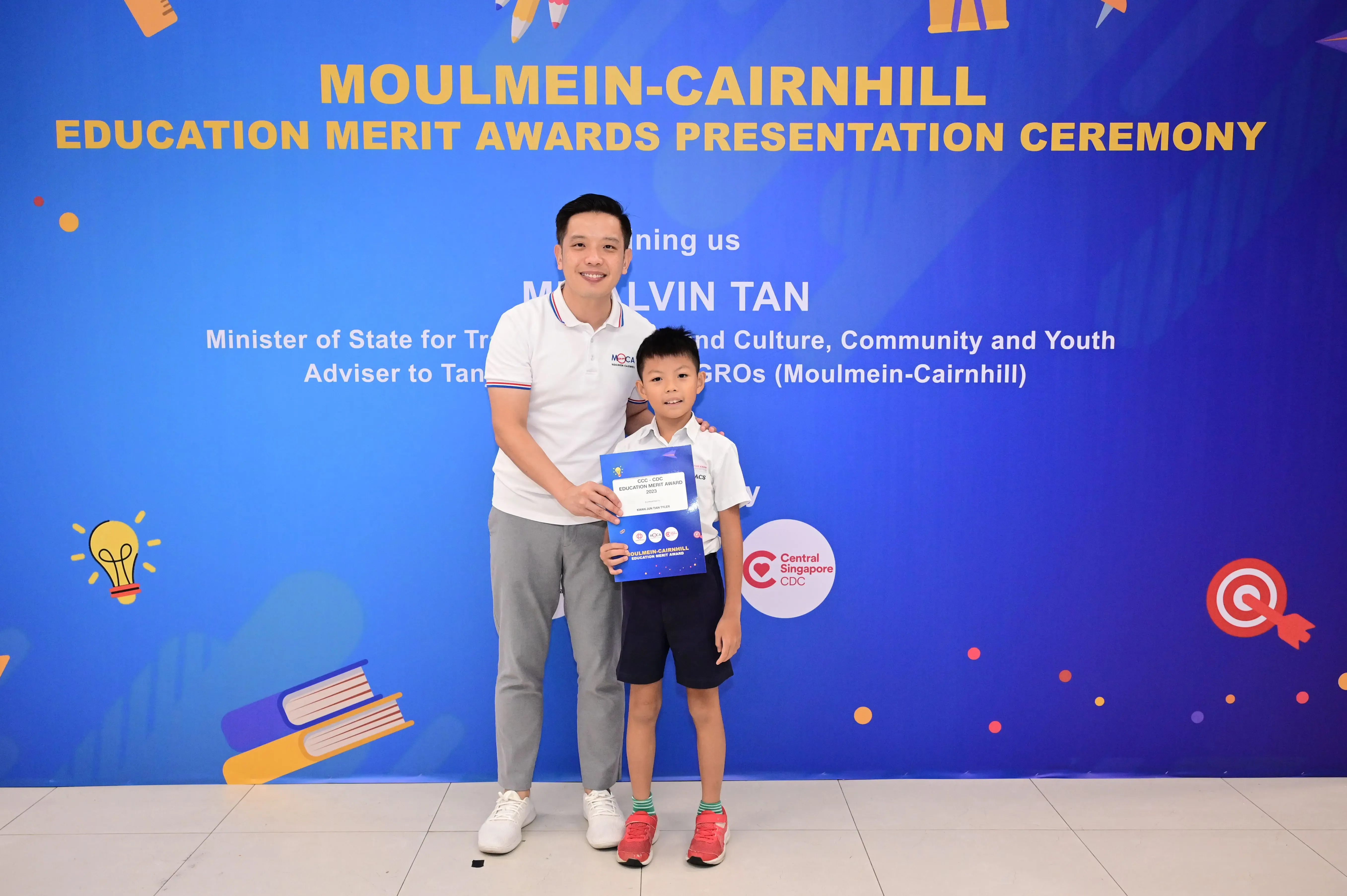 A man and a young boy pose together holding a certificate in front of a Moulmein-Cairnhill Education Merit Awards banner.