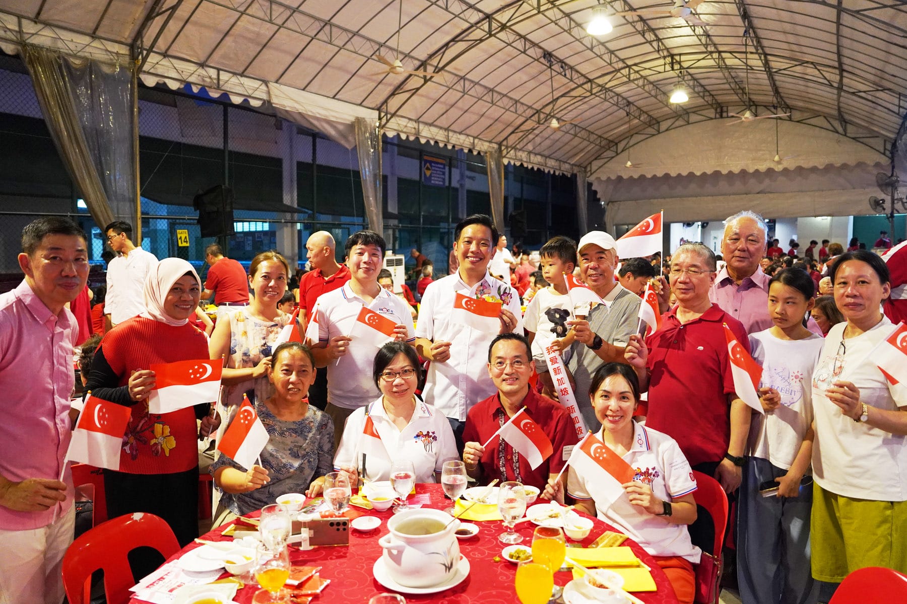 A large group of smiling people holding small Singapore flags at a festive outdoor dinner under a covered tent.