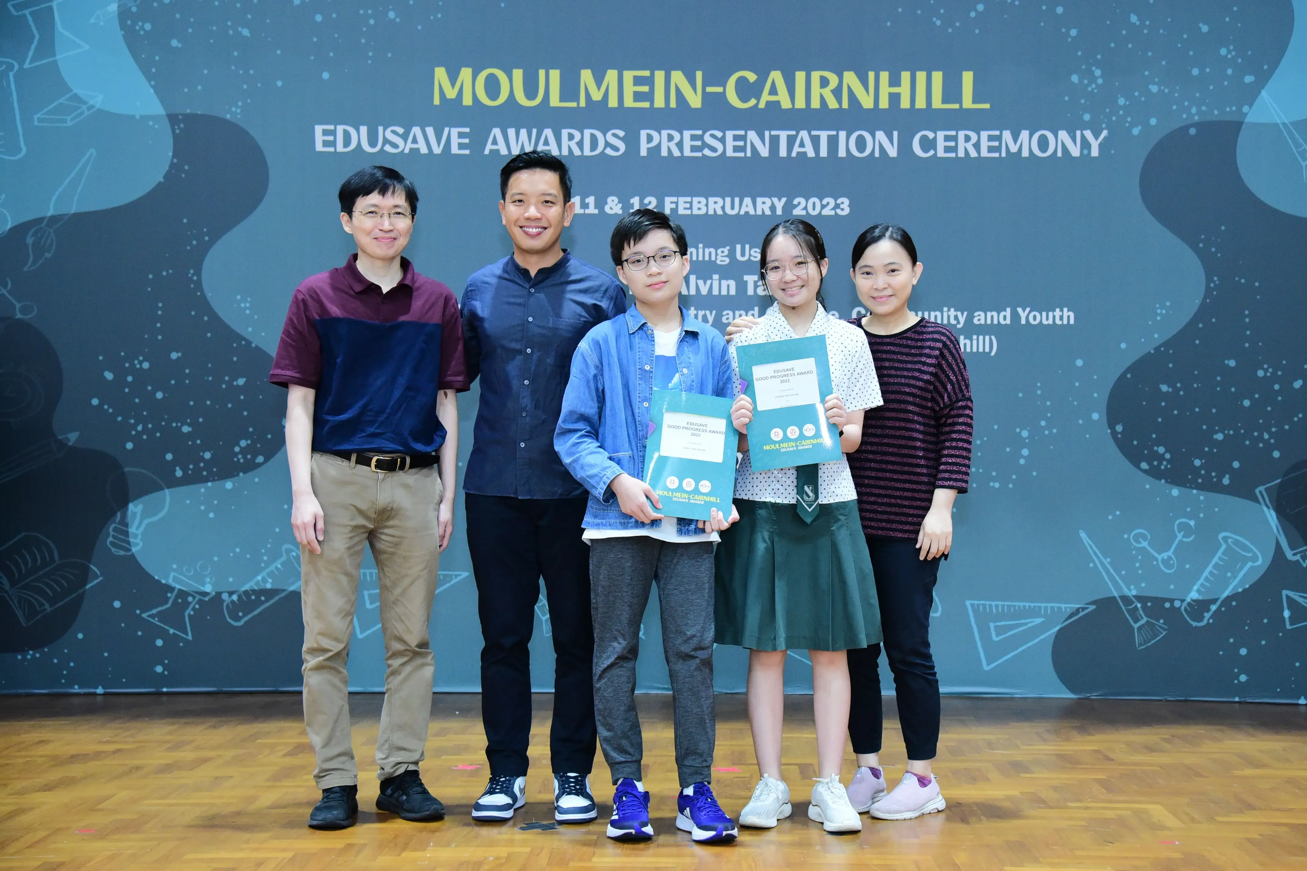 Five people pose at the Moulmein-Cairnhill Edusave Awards Presentation Ceremony, two children holding award folders.
