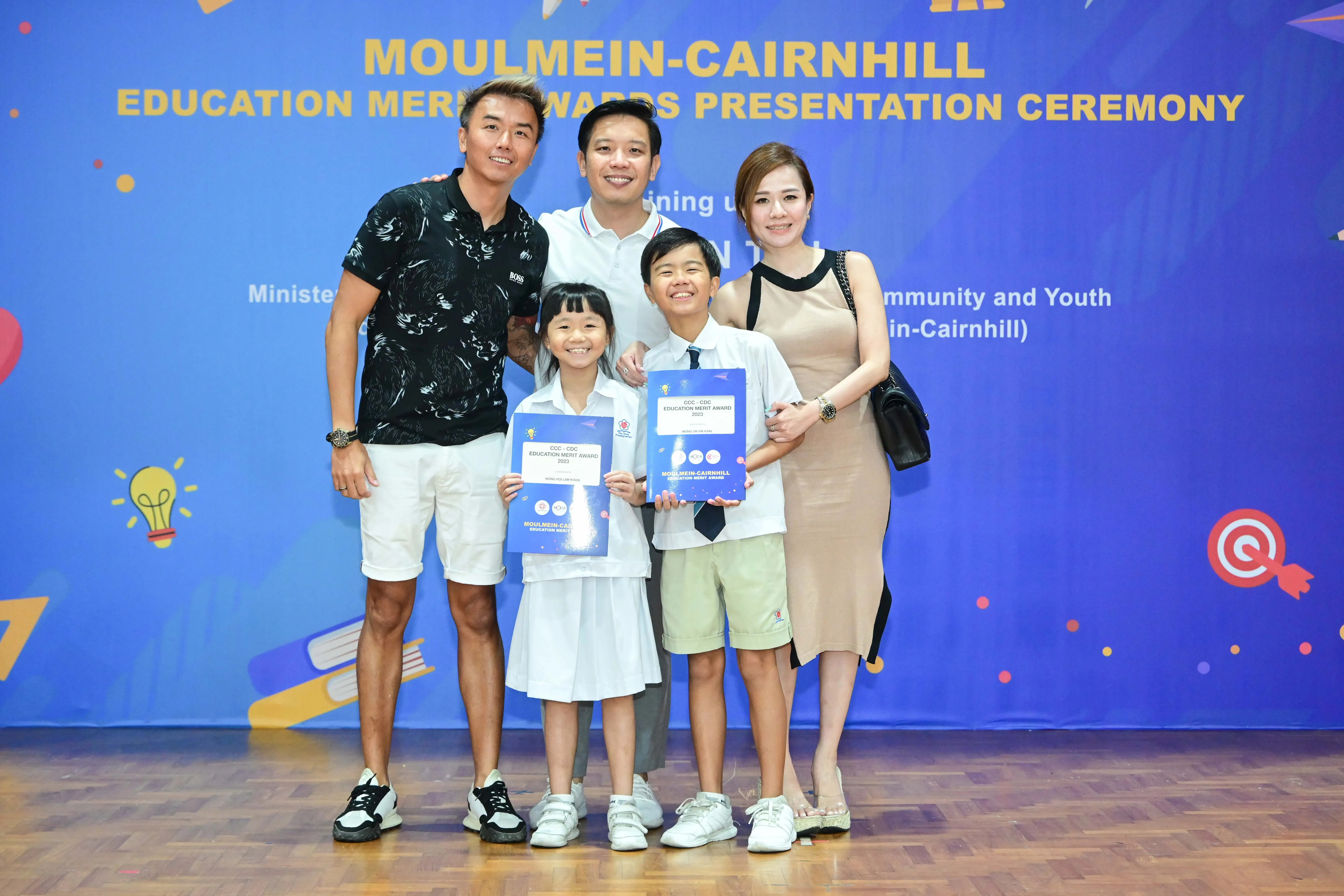 Five people pose before a Moulmein-Cairnhill Education Merit Awards banner, two children holding award certificates.