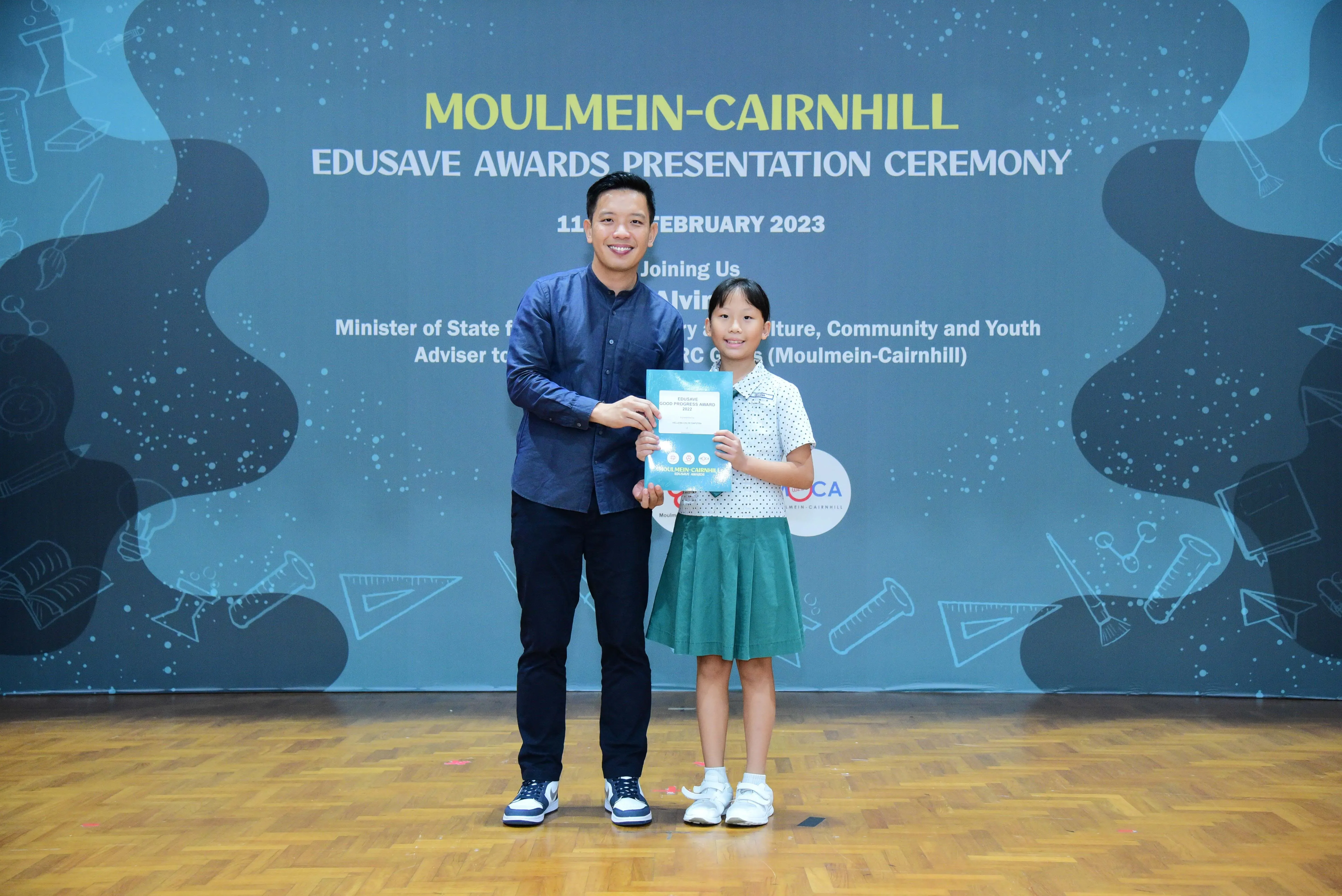 A man and a young girl hold a certificate together on stage at the Moulmein-Cairnhill Edusave Awards Ceremony 2023.