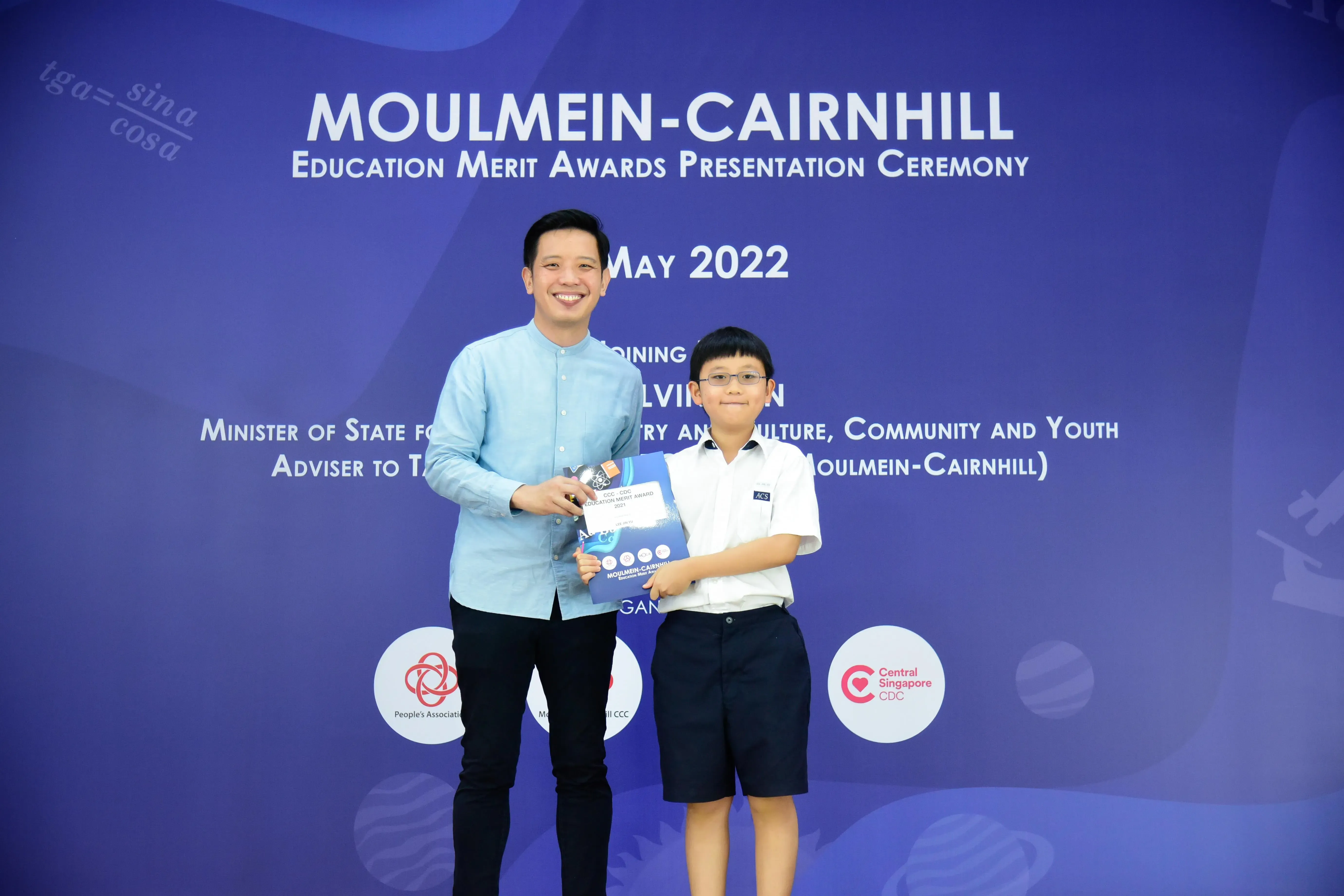 A man and a young boy in school uniform hold an award together at the Moulmein-Cairnhill Education Merit Awards, May 2022.