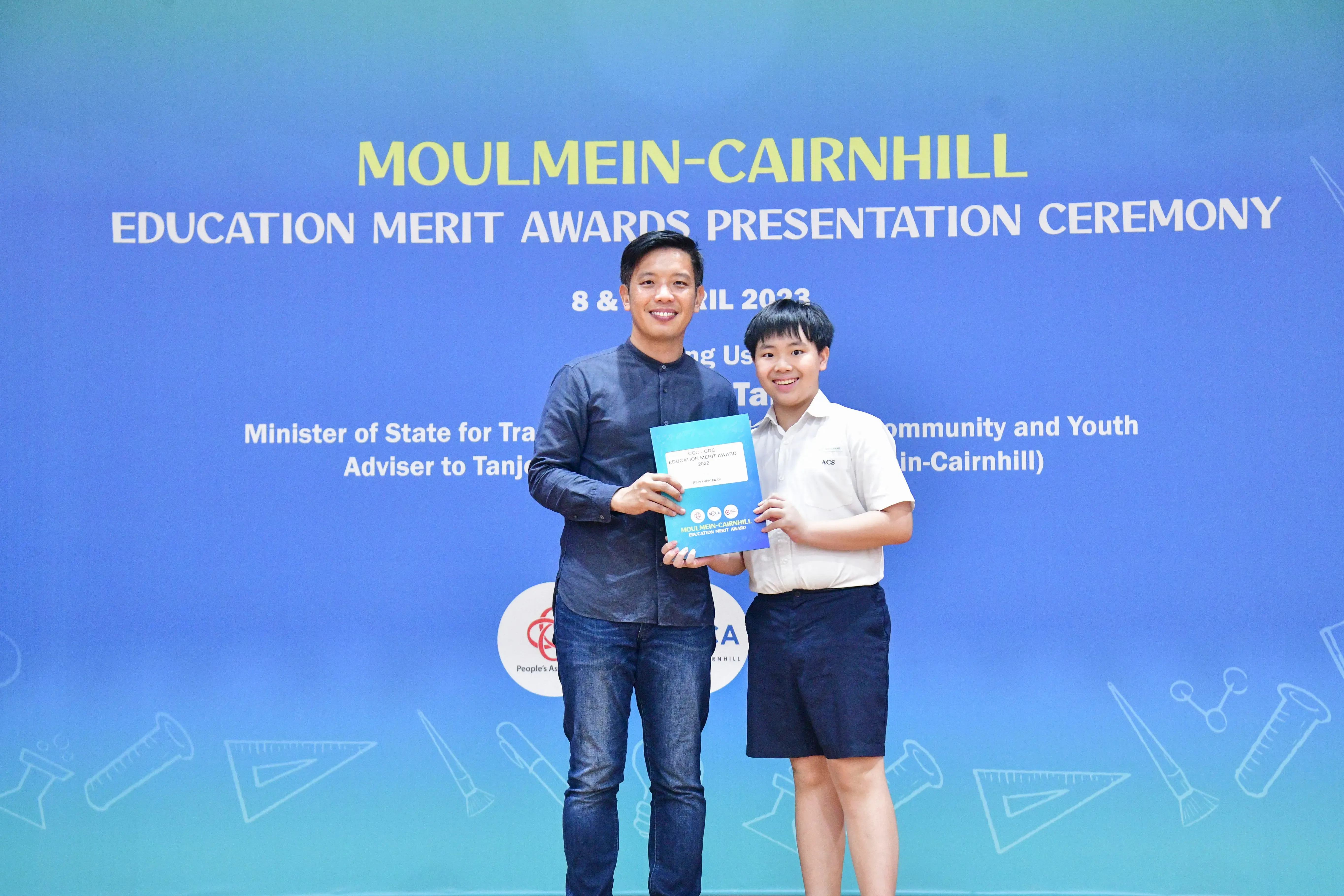 Two people hold a certificate together in front of a Moulmein-Cairnhill Education Merit Awards Ceremony banner.