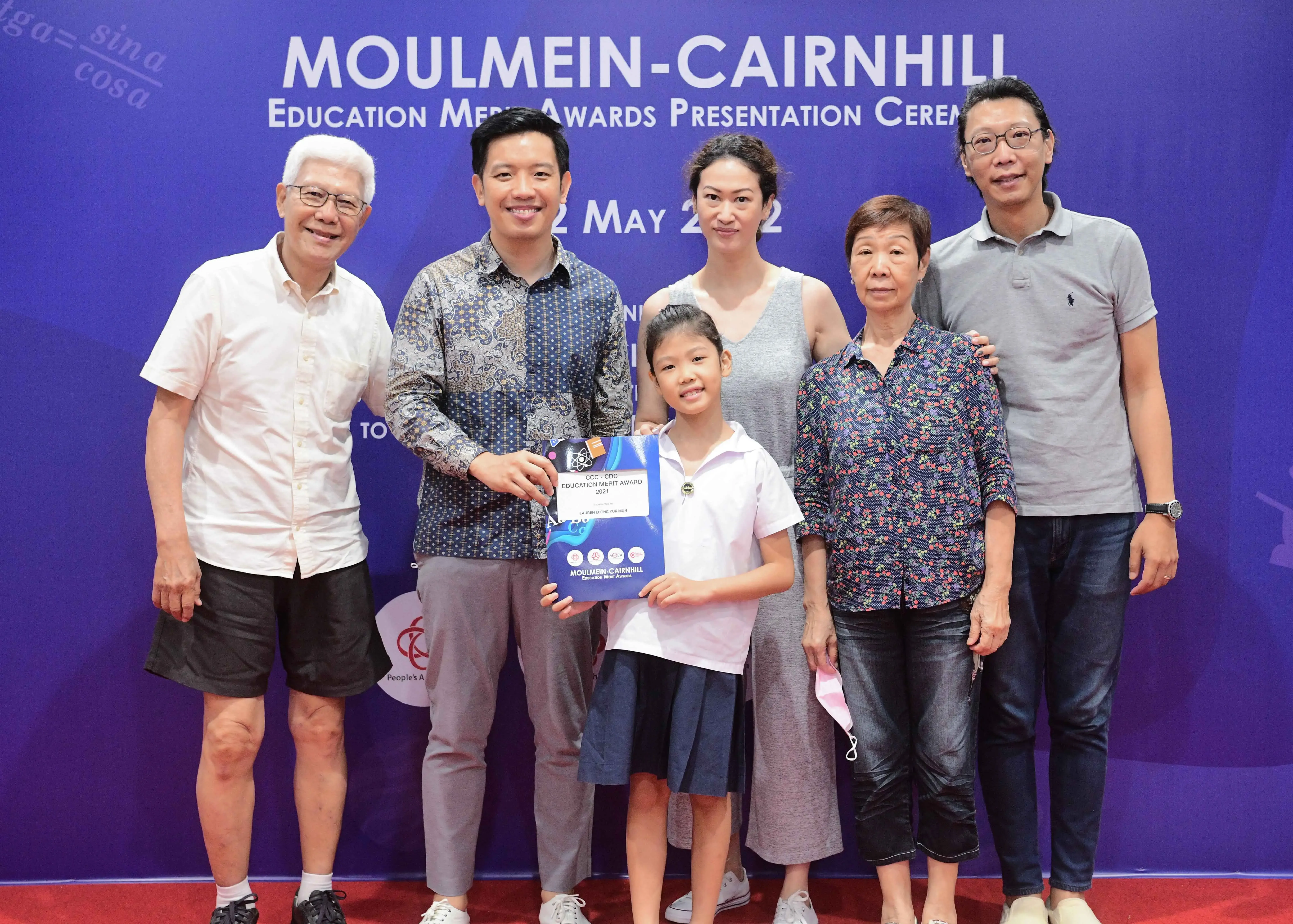Six people pose at the Moulmein-Cairnhill Education Merit Awards ceremony, with a young girl holding an award certificate.