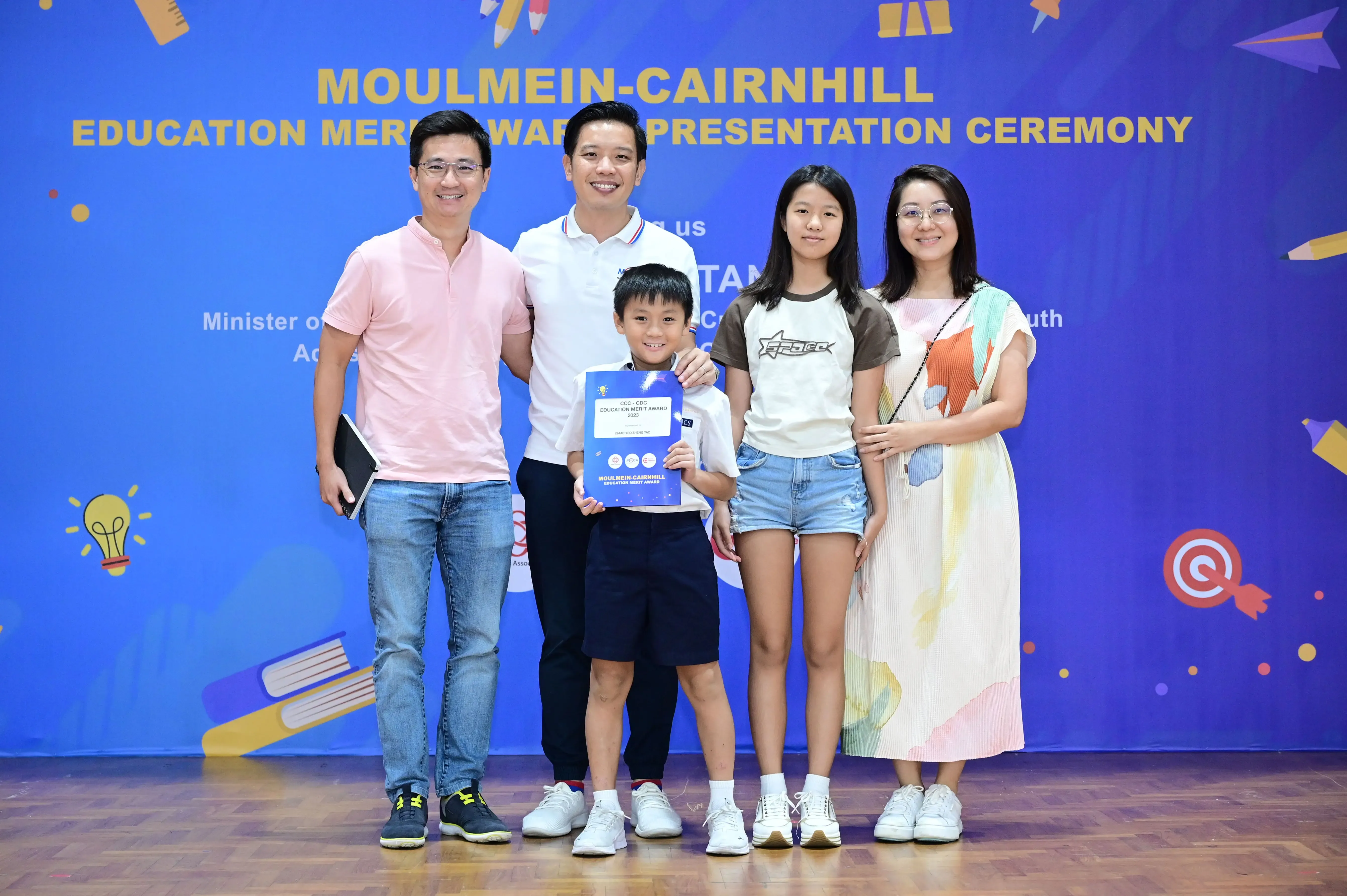 Five people pose at the Moulmein-Cairnhill Education Merit Award Presentation Ceremony, with a boy holding a certificate.