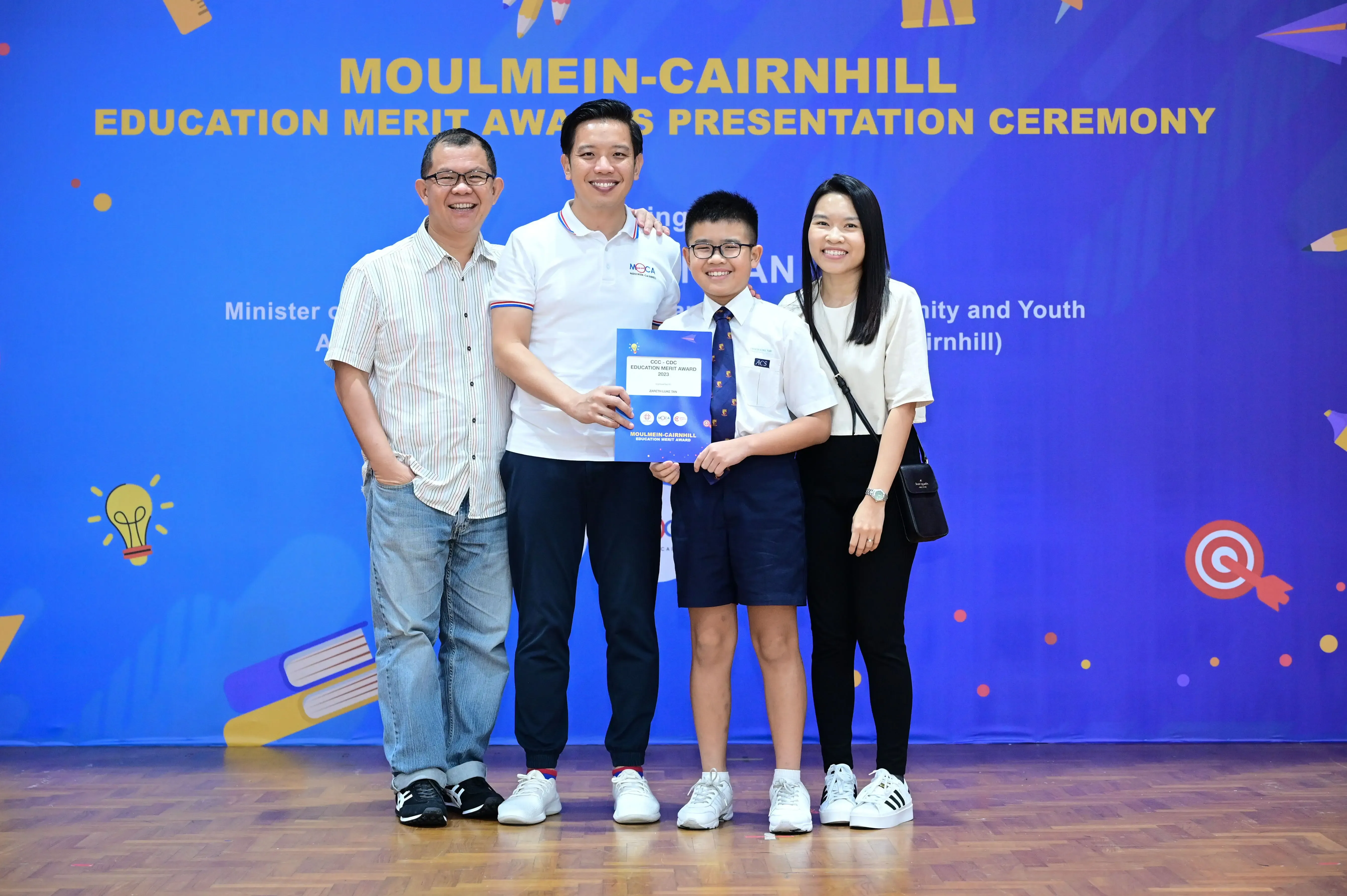 Four people pose smiling at the Moulmein-Cairnhill Education Merit Awards Presentation Ceremony, with a boy holding a certificate.