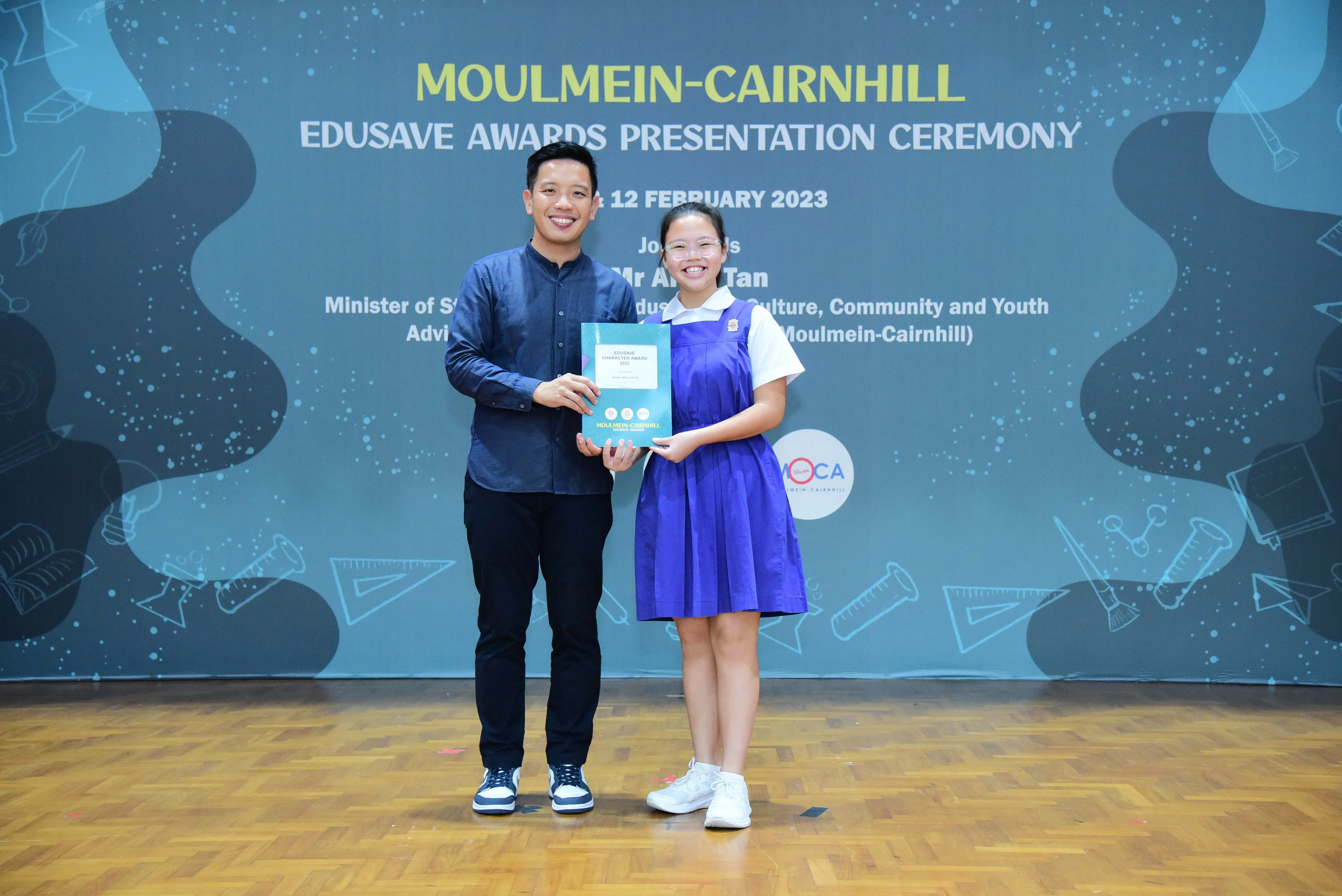 A man and a young girl in a blue school uniform hold a certificate at the Moulmein-Cairnhill Edusave Awards Ceremony.