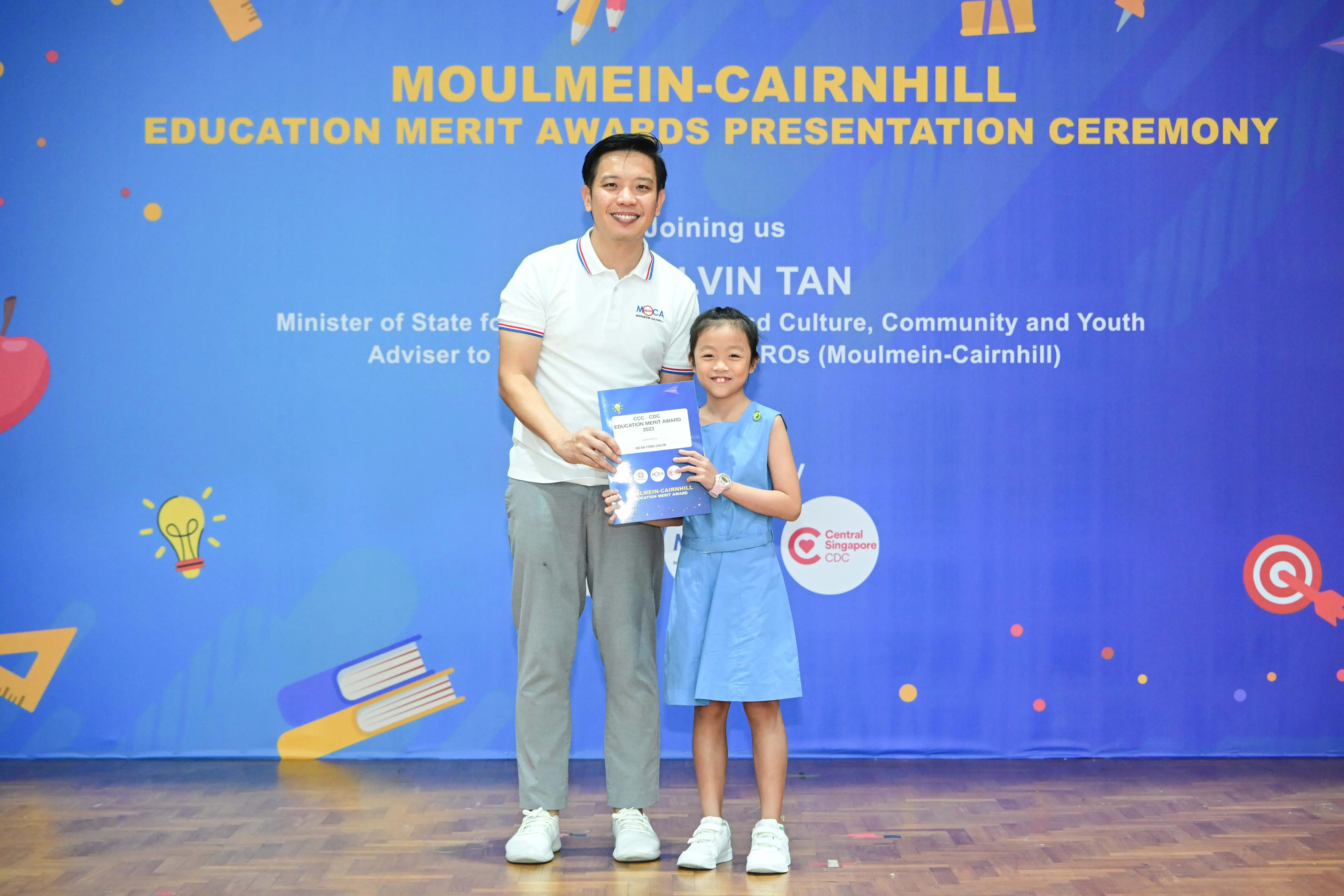 A man and a young girl hold a certificate in front of a Moulmein-Cairnhill Education Merit Awards banner.
