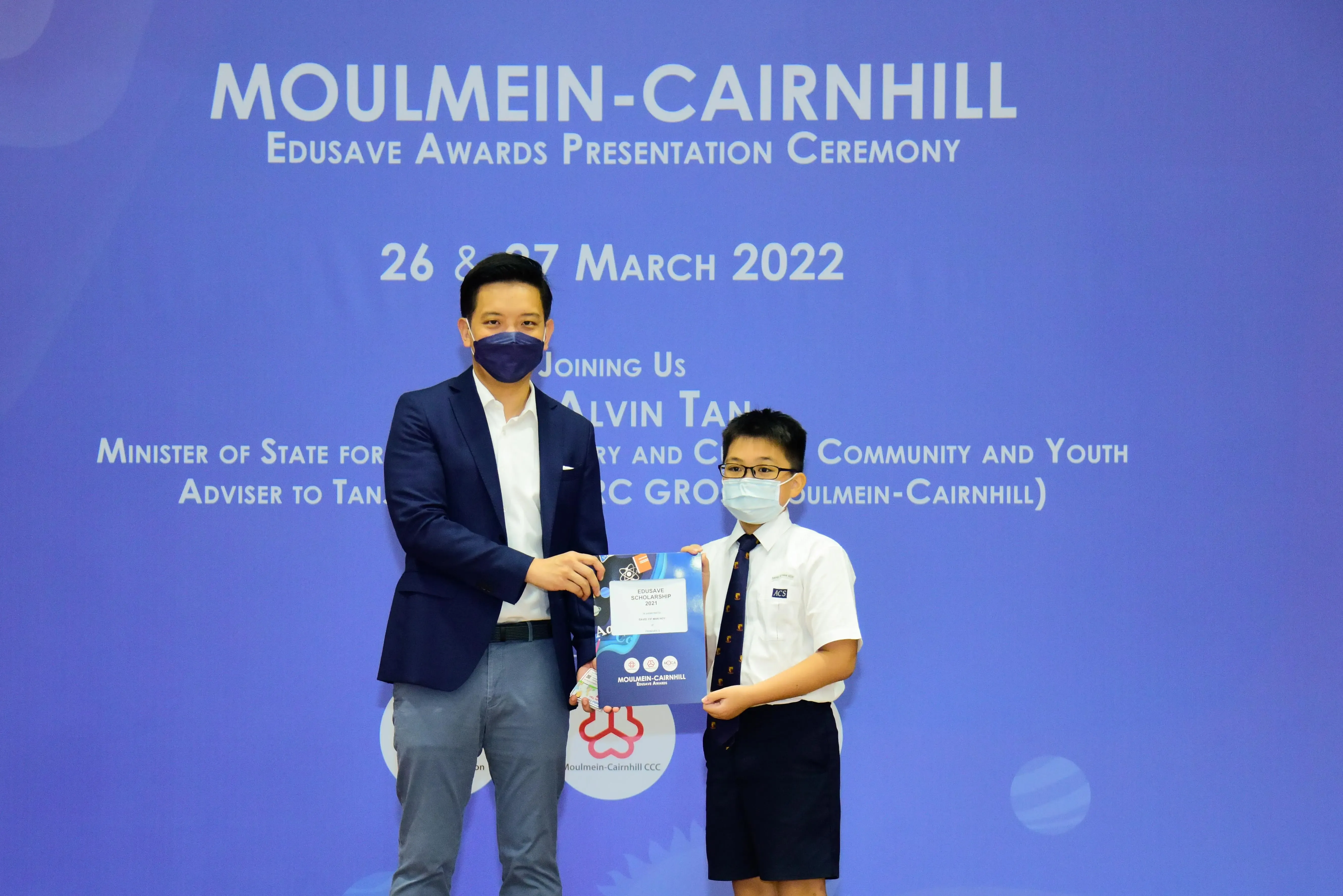 An adult and a young student in uniform hold an award folder at the Moulmein-Cairnhill Edusave Awards Ceremony, March 2022.