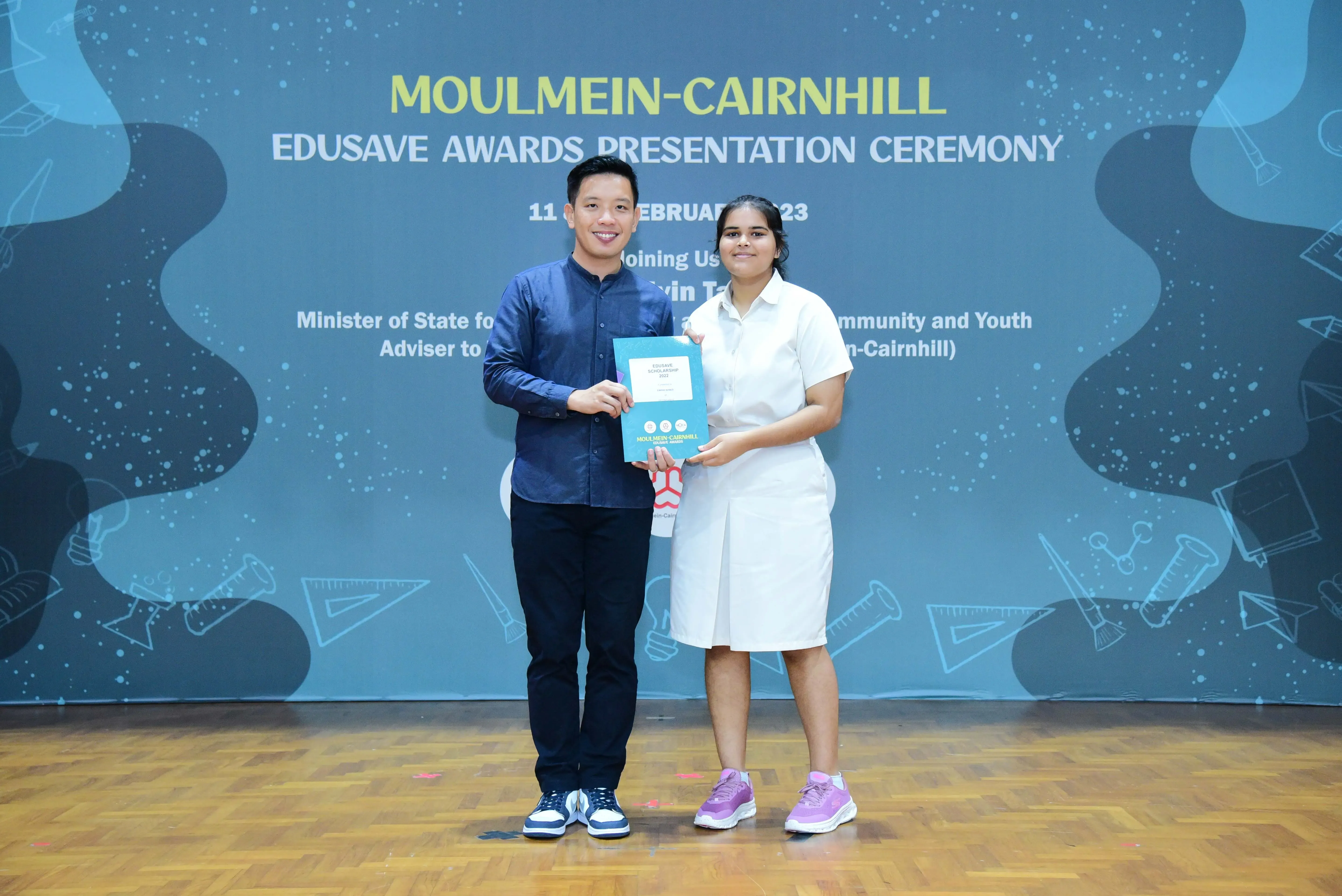 Two people hold a certificate together on stage at the Moulmein-Cairnhill Edusave Awards Presentation Ceremony.