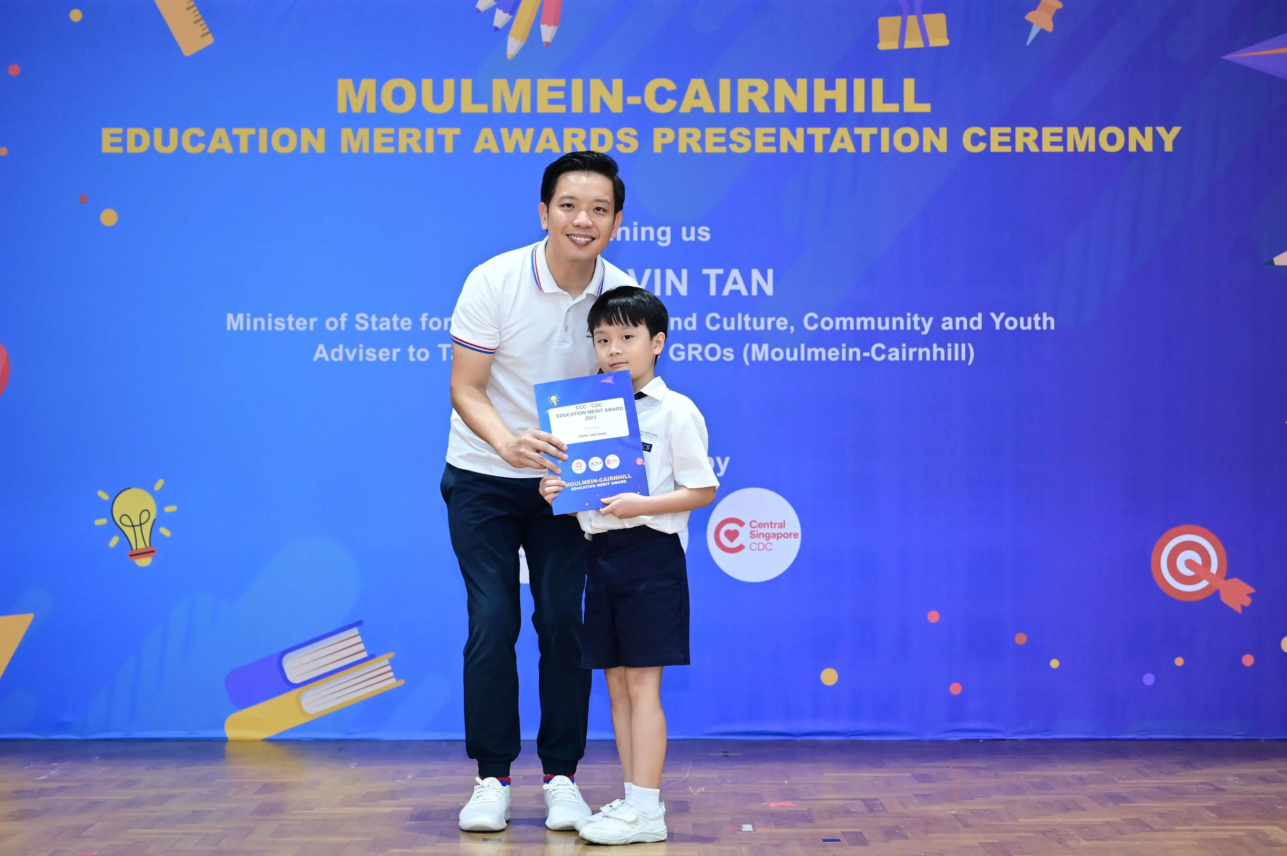 A man and a young boy hold a certificate in front of the Moulmein-Cairnhill Education Merit Awards banner.
