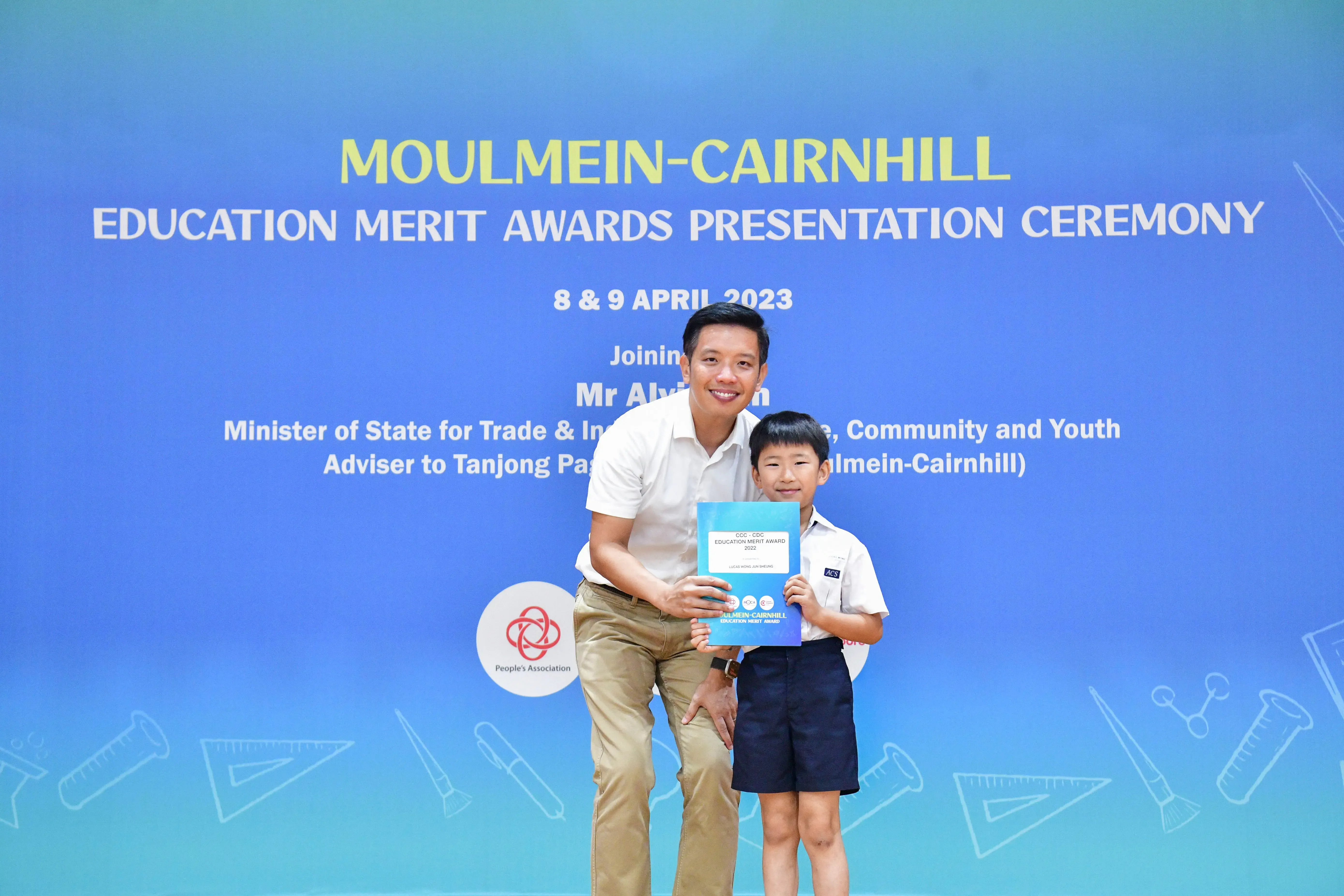 A man and a young boy hold a certificate together in front of a Moulmein-Cairnhill Education Merit Awards banner.