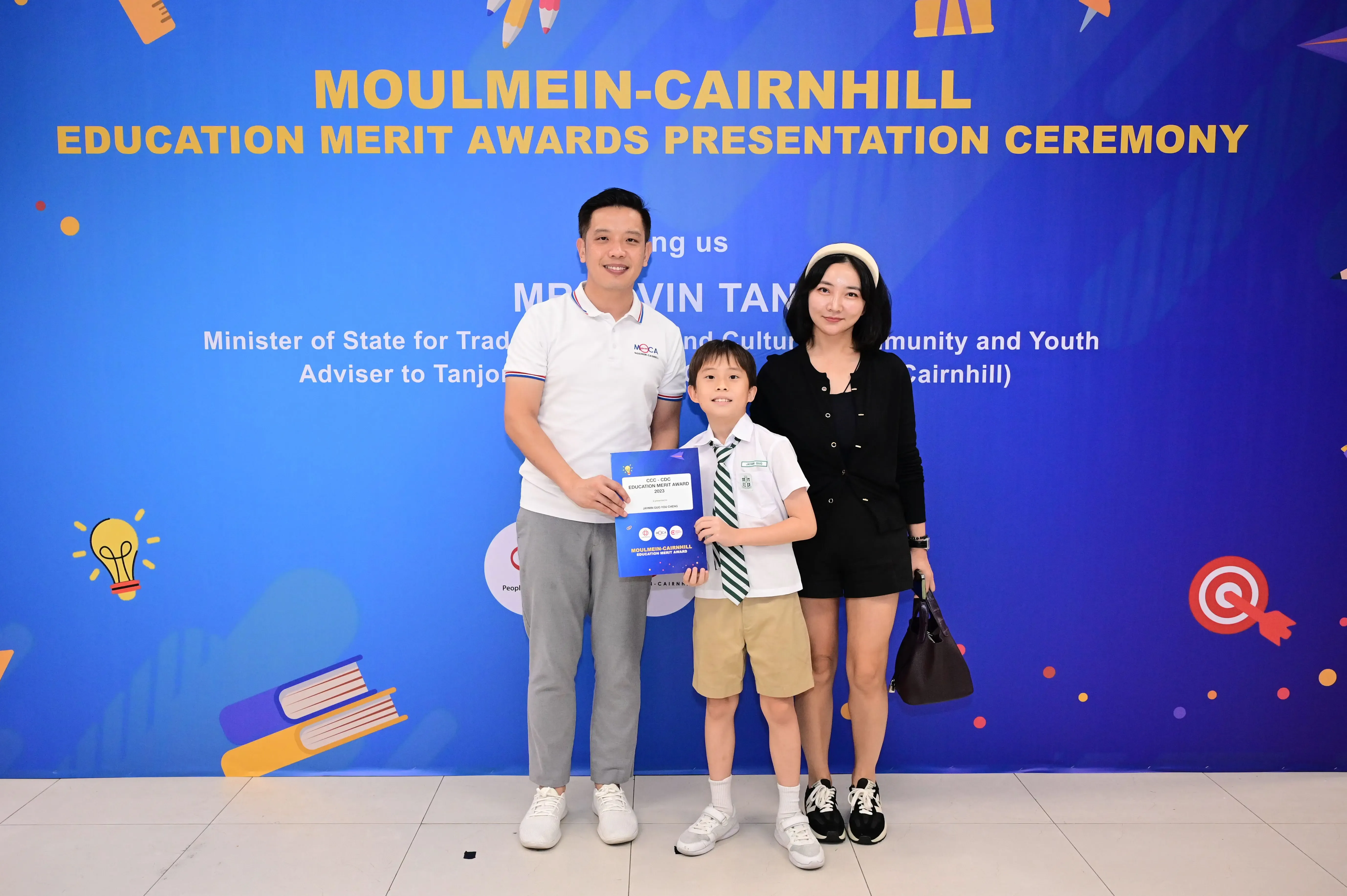A man, woman, and young boy hold a certificate in front of a Moulmein-Cairnhill Education Merit Awards banner.