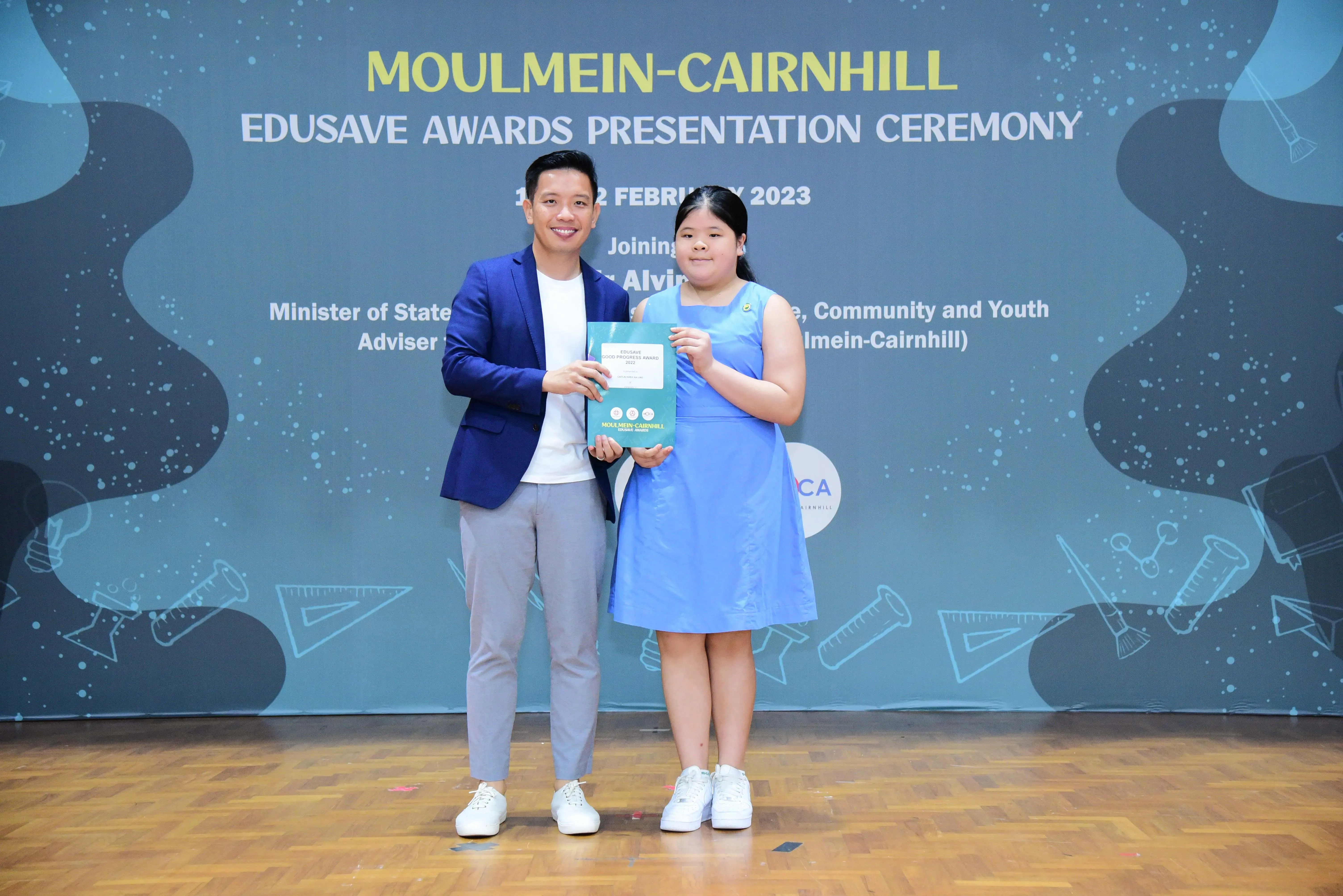 A man in a blue blazer and a girl in a blue dress hold a certificate at the Moulmein-Cairnhill Edusave Awards Ceremony 2023.