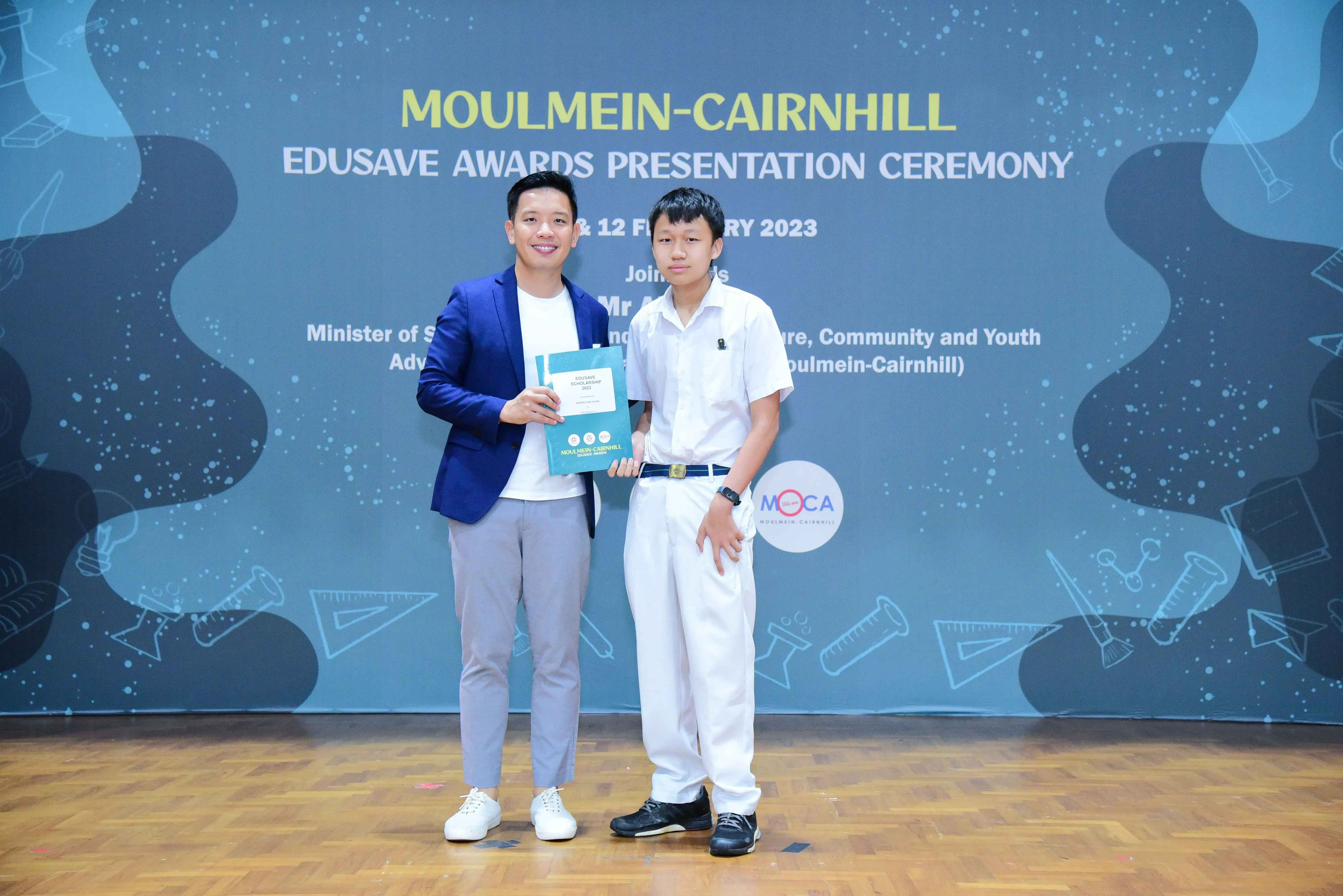 Two people pose at the Moulmein-Cairnhill Edusave Awards Presentation Ceremony 2023, one holding an award folder.