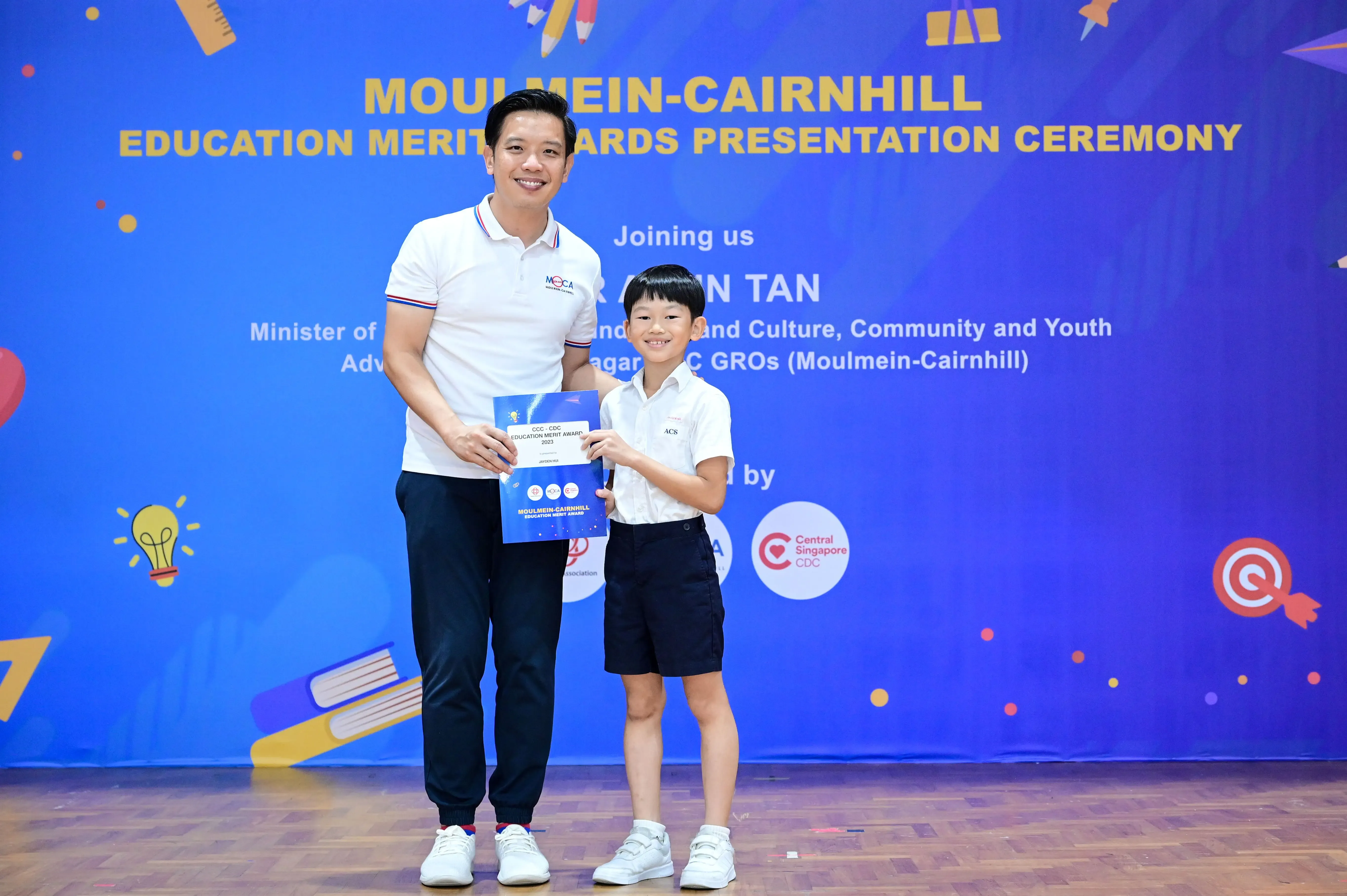 Man and young boy hold a blue certificate together at Moulmein-Cairnhill Education Merit Awards ceremony.