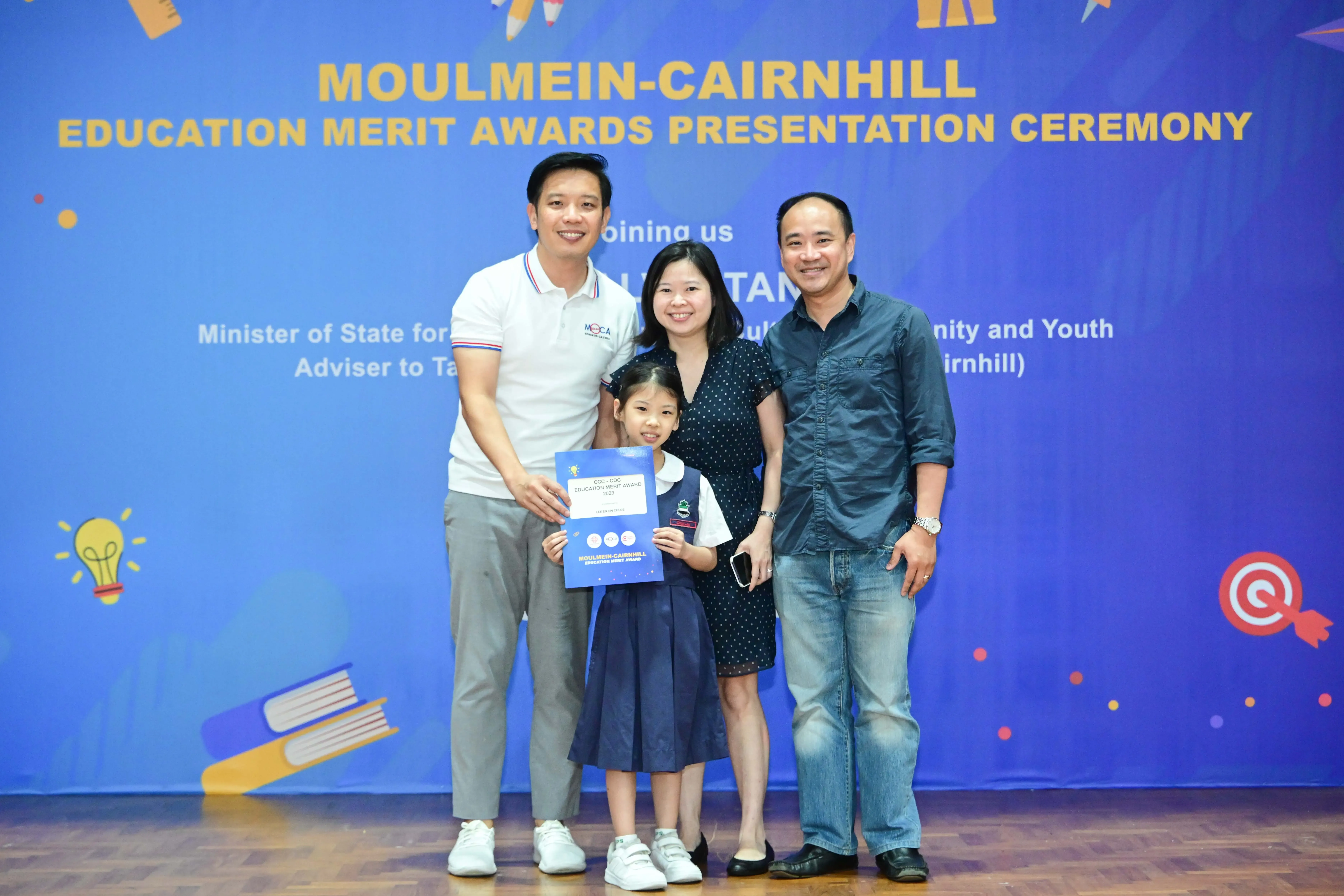 Three adults and a young girl holding a certificate pose before a Moulmein-Cairnhill Education Merit Awards banner.