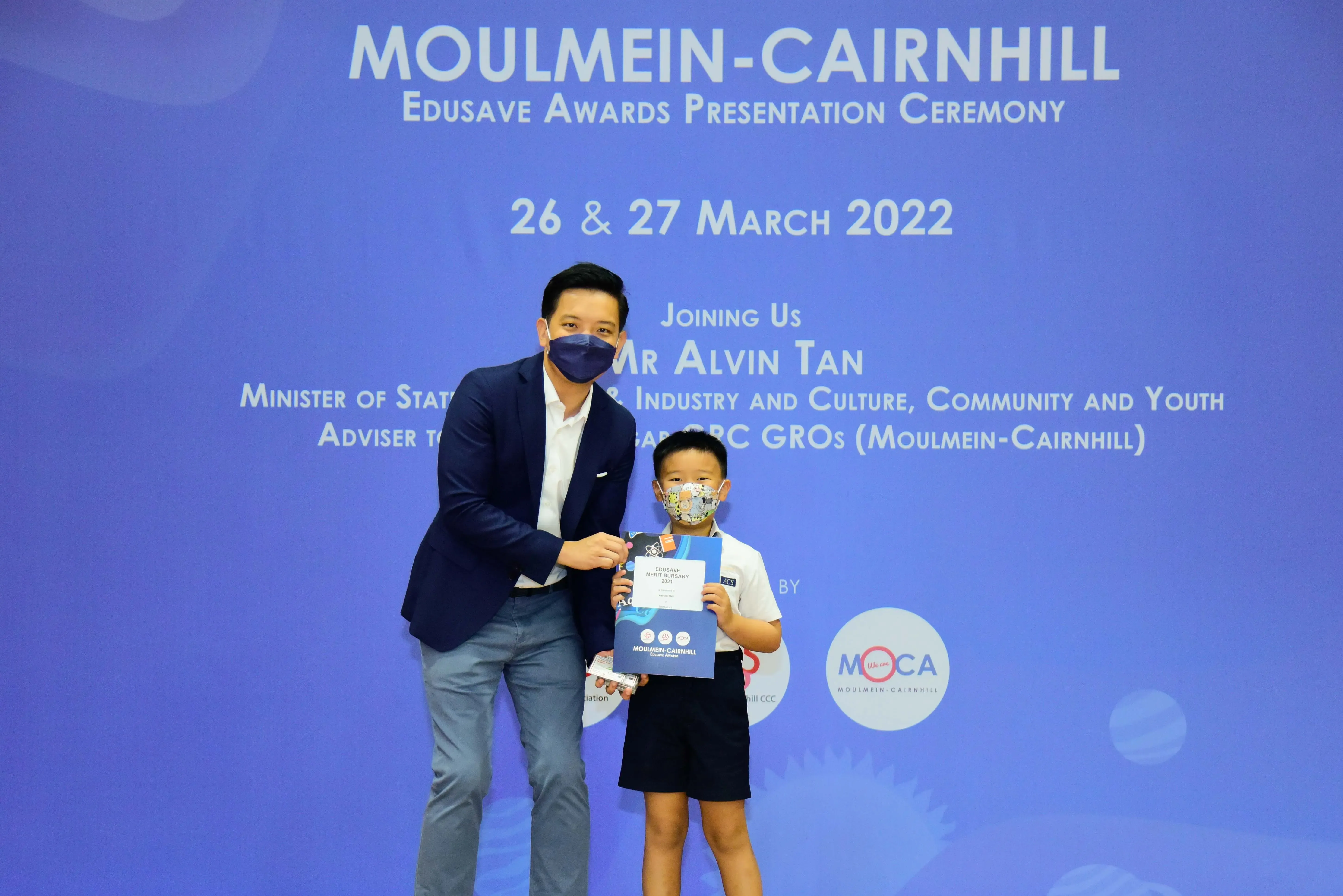 An adult and a young child in masks hold a certificate at the Moulmein-Cairnhill Edusave Awards Ceremony, March 2022.